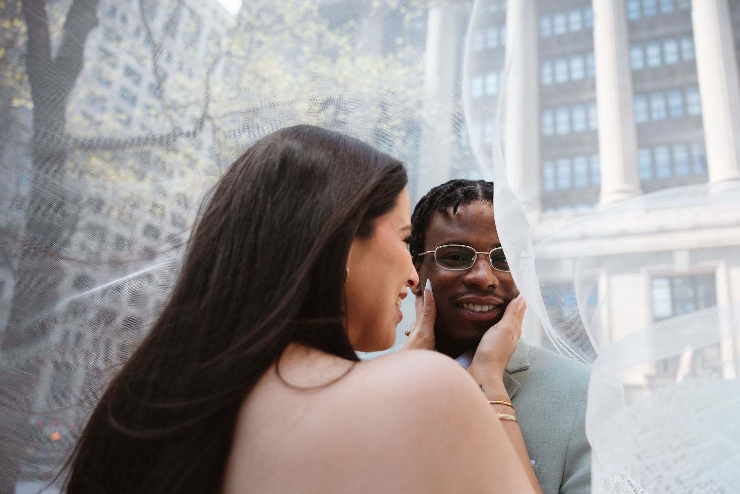 Bride and groom share intimate moment under wedding veil with city buildings in background.