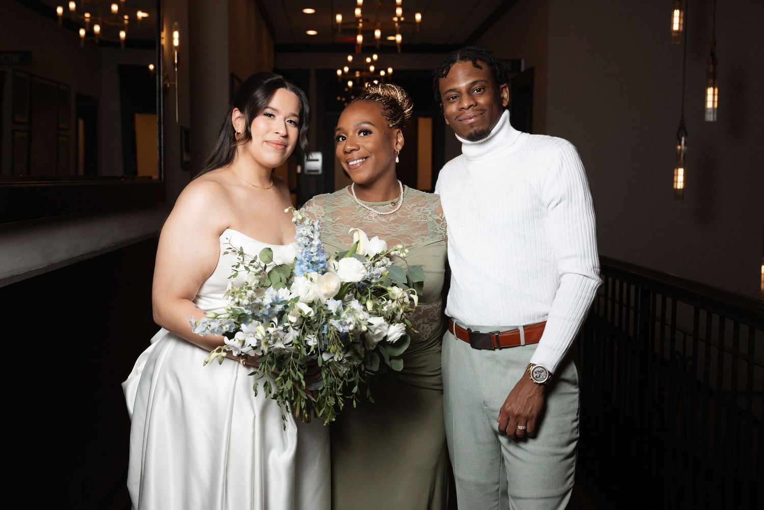 Bride holding blue and white bouquet poses with two guests at elegant indoor wedding reception.