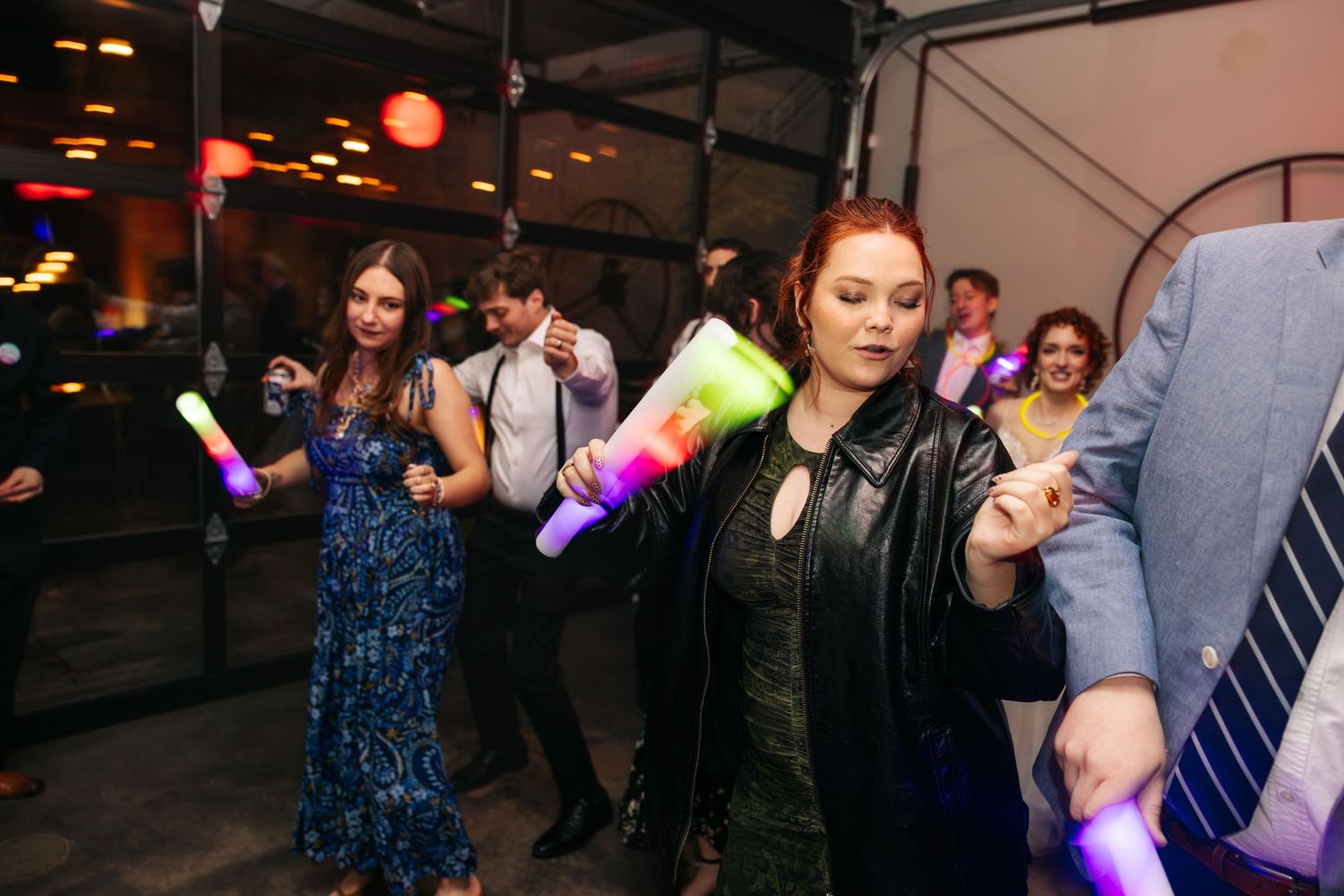 Bride in a leather jacket dances energetically with guests holding glowing foam sticks at a lively wedding reception.