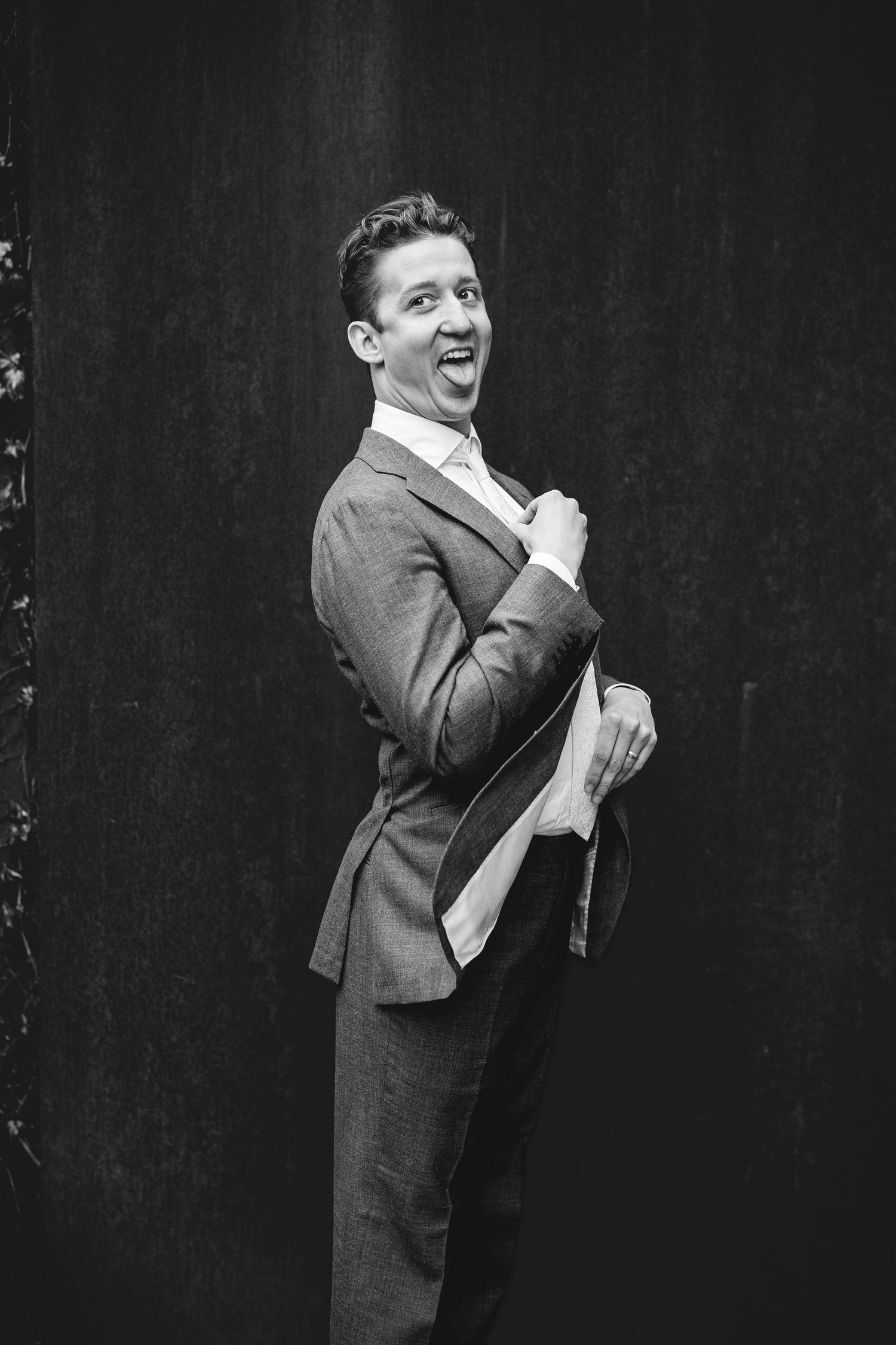 Black and white portrait of groom in suit laughing while adjusting tie against dark background.
