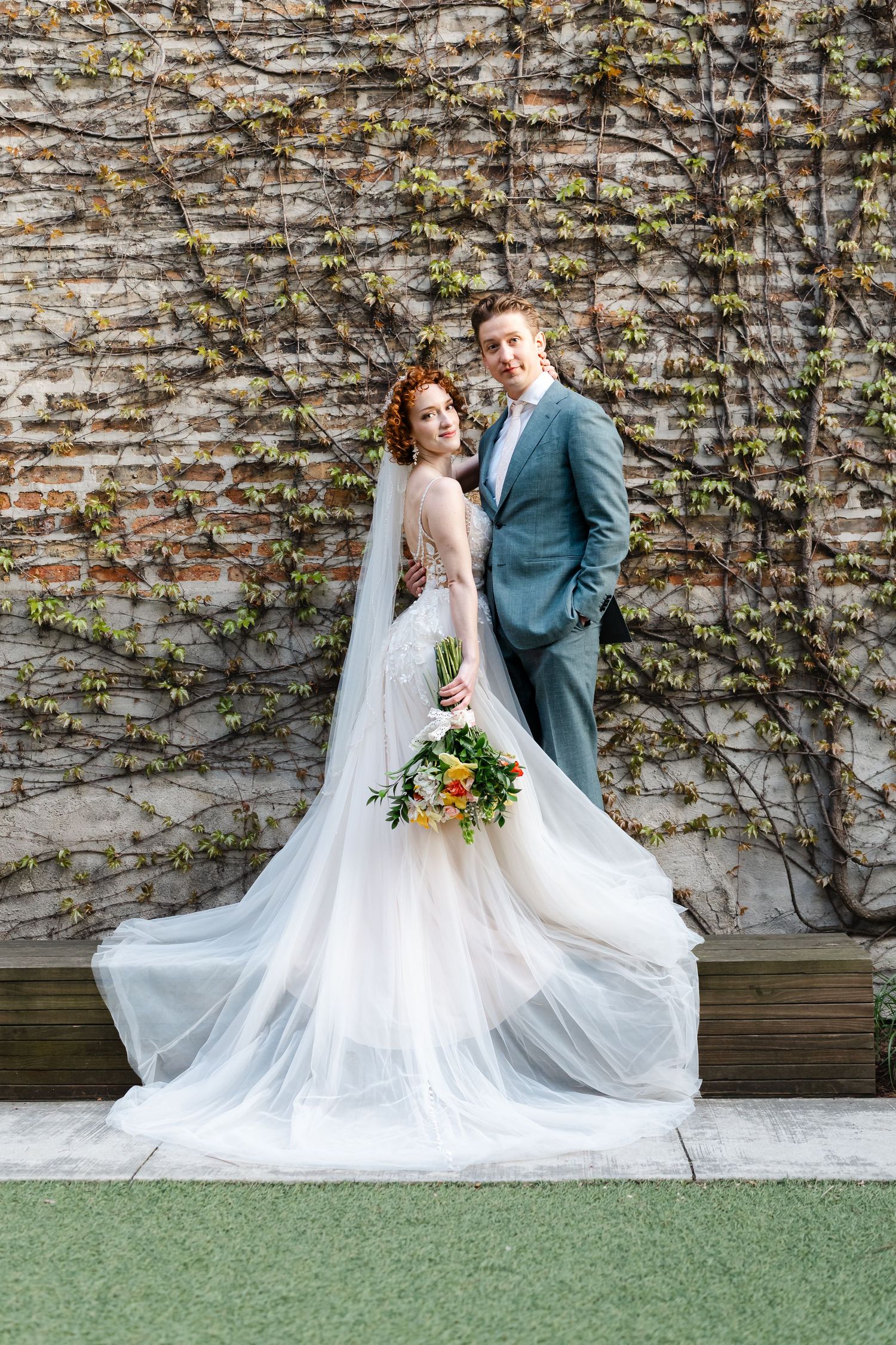 Bride in flowing white gown and groom in teal suit pose before ivy-covered brick wall.
