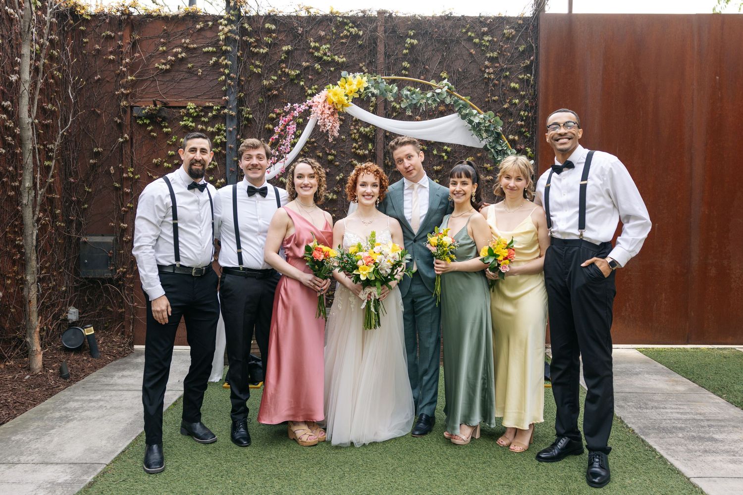 Wedding party of seven poses outdoors under floral arch with colorful bouquets in coordinated attire.