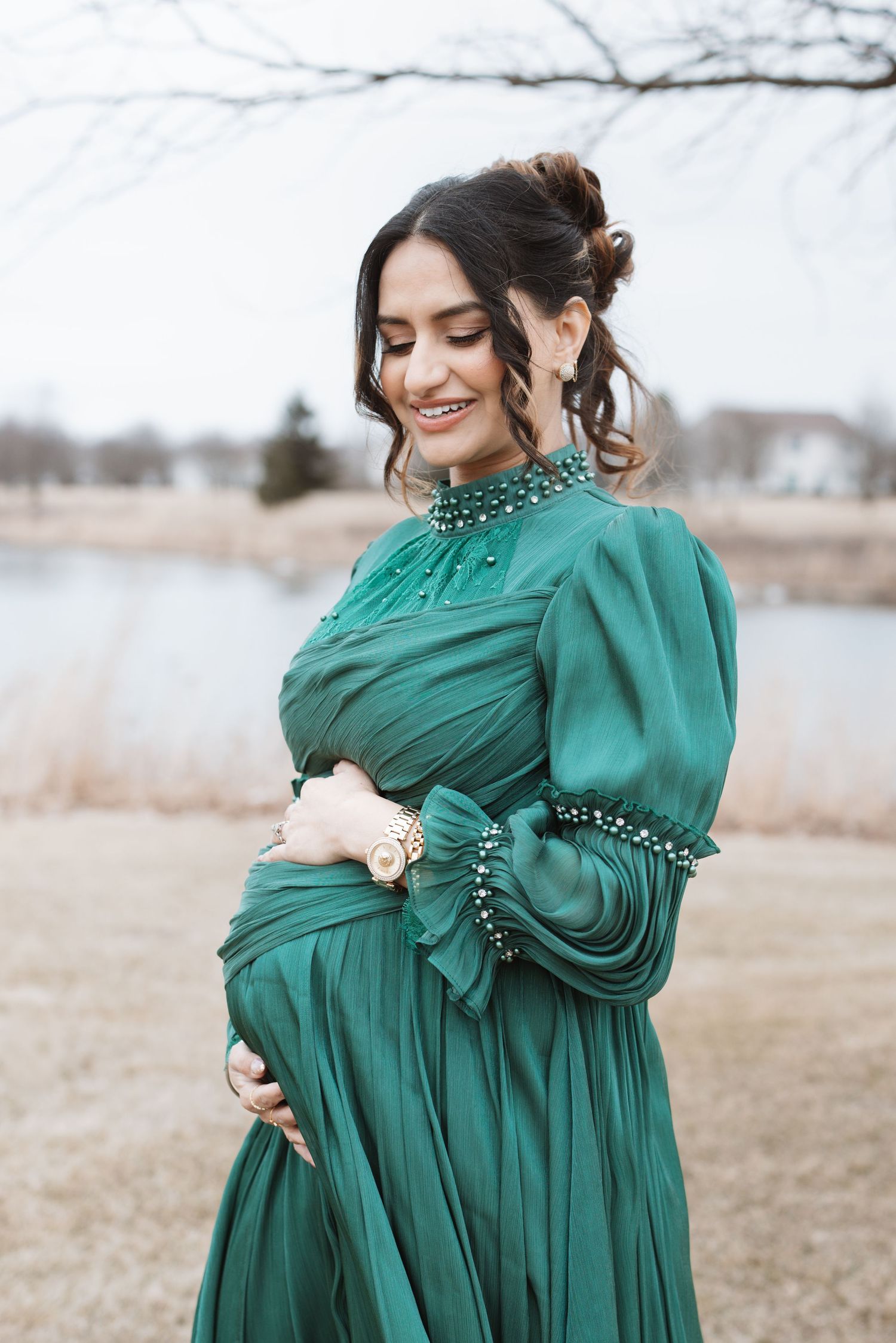Pregnant woman in emerald green gown smiling while holding baby bump outdoors near a lake.