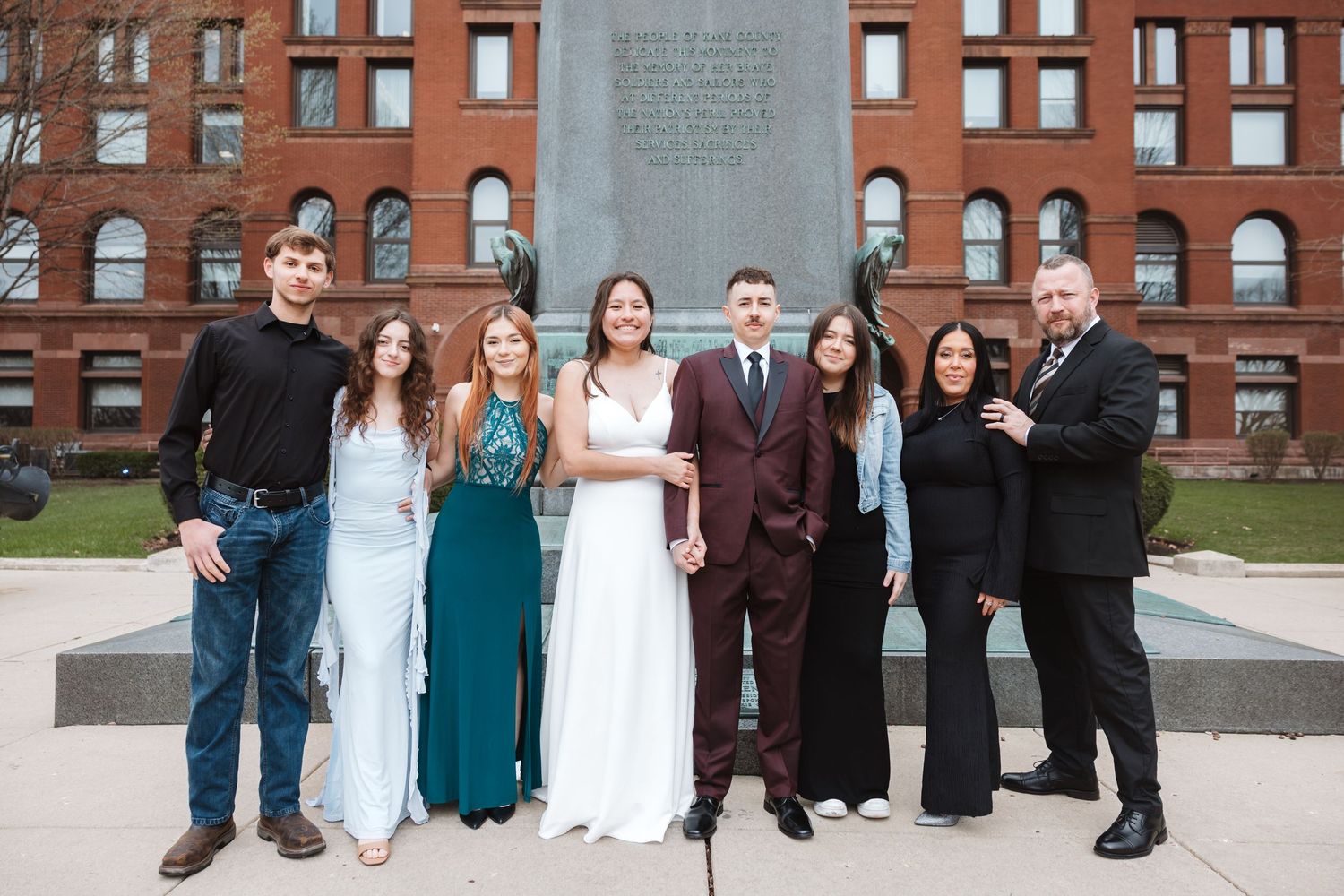 Group of eight people posing outdoors in formal attire near a monument with a brick building backdrop.