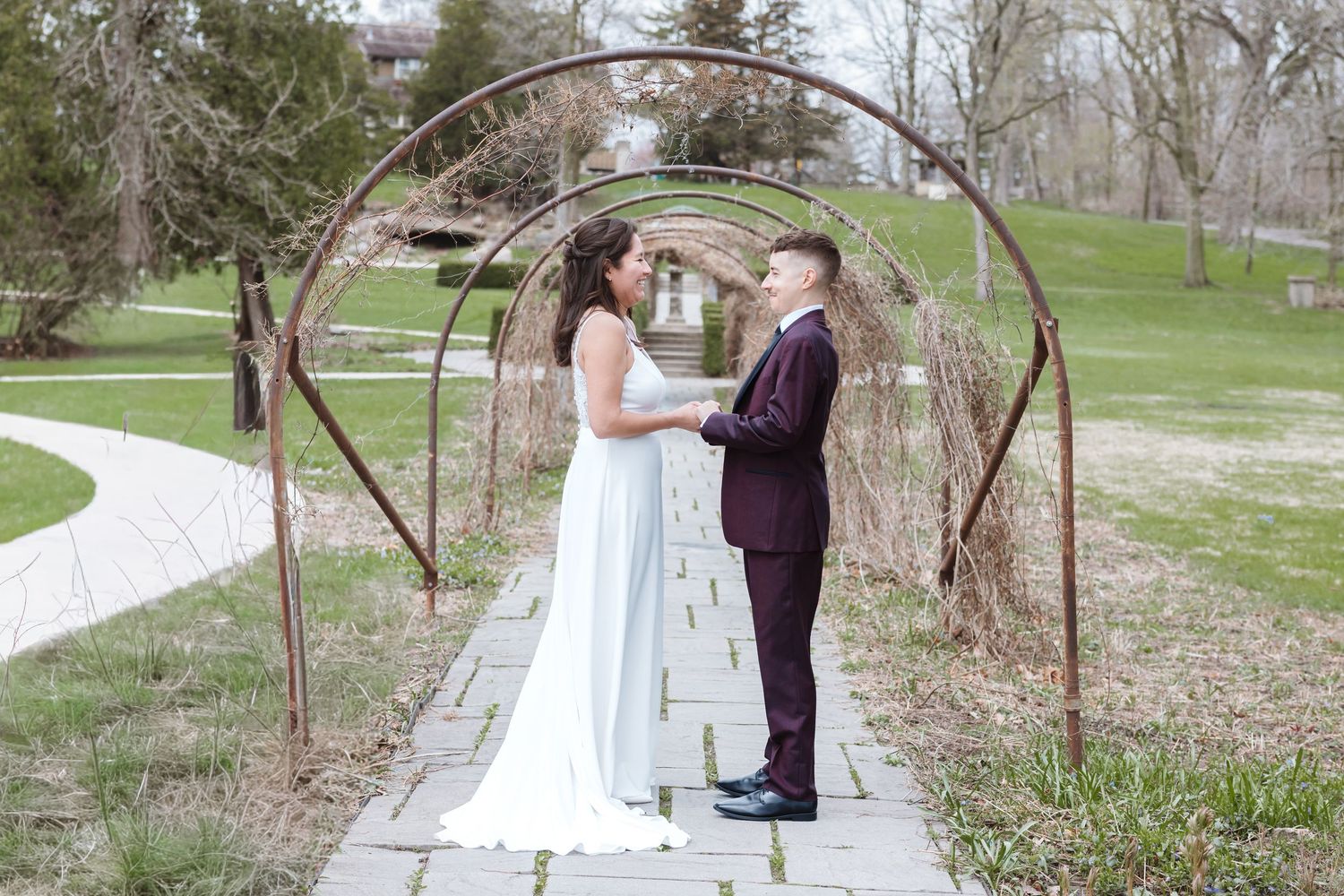 Bride and groom hold hands under rustic iron arch on stone path in lush green park setting.