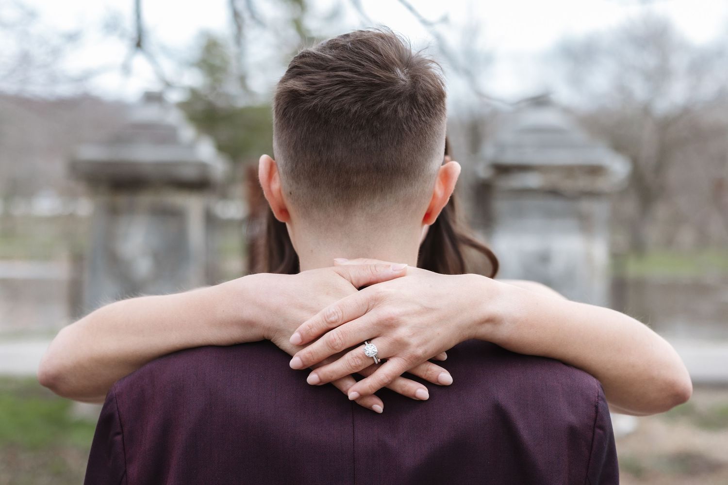 Couple embracing from behind, woman's hand with engagement ring resting on man's shoulder outdoors.
