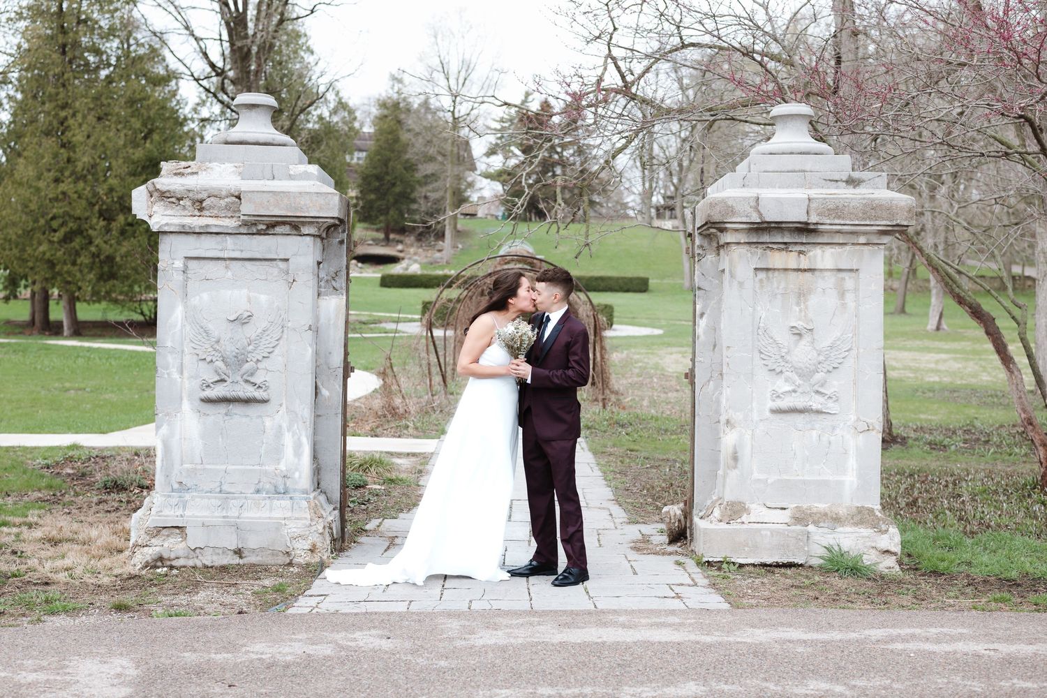 Bride and groom kissing between stone gate pillars in a lush green park setting on their wedding day.