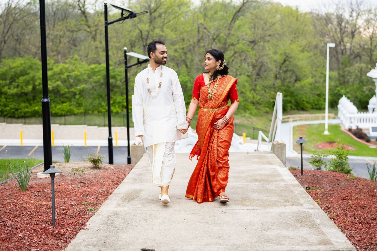 Indian bride and groom in traditional attire smiling while walking hand-in-hand outdoors.