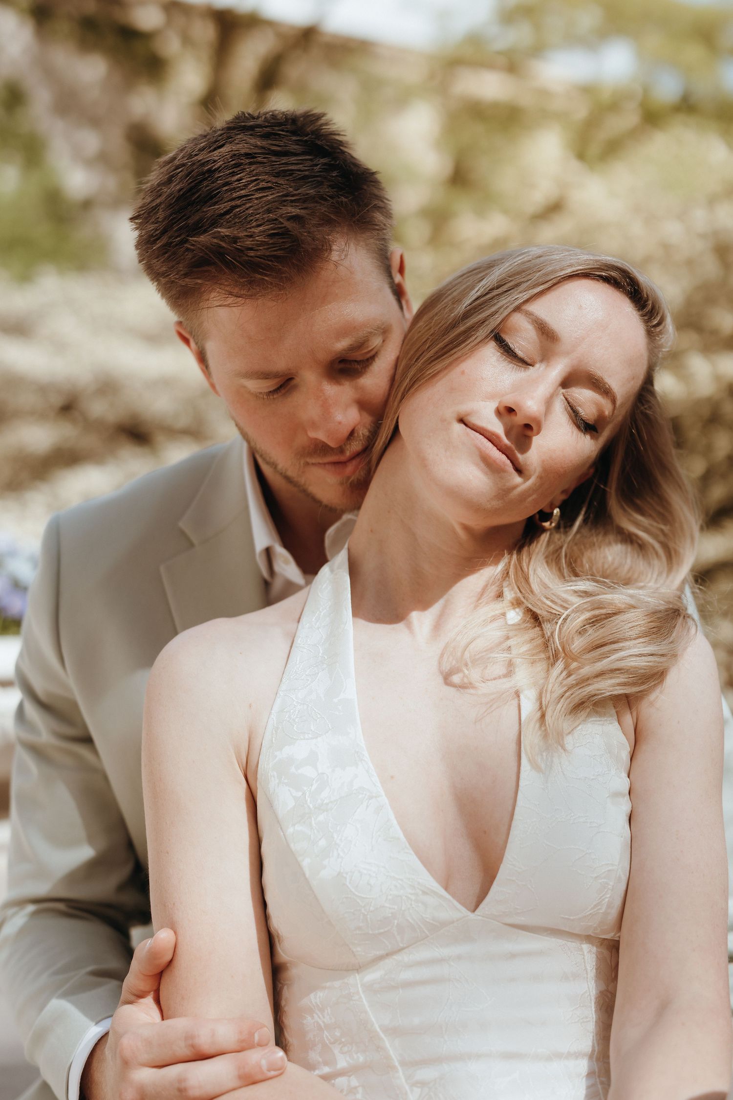 Groom in gray suit embraces bride in white lace dress from behind as she tilts her head back.