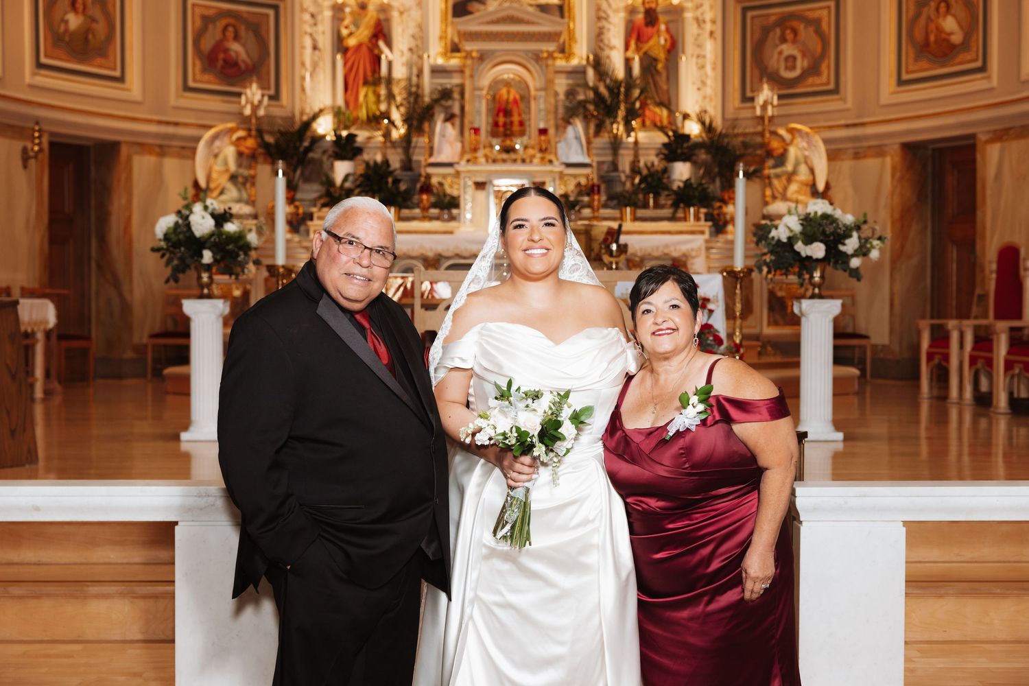 Bride in white satin gown poses with parents inside a beautiful decorated church after wedding ceremony.