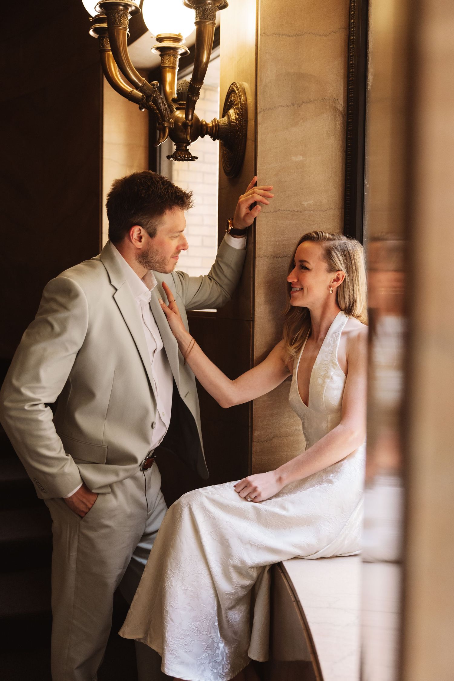 Bride in white dress and groom in light suit share a playful moment by an ornate wall sconce.
