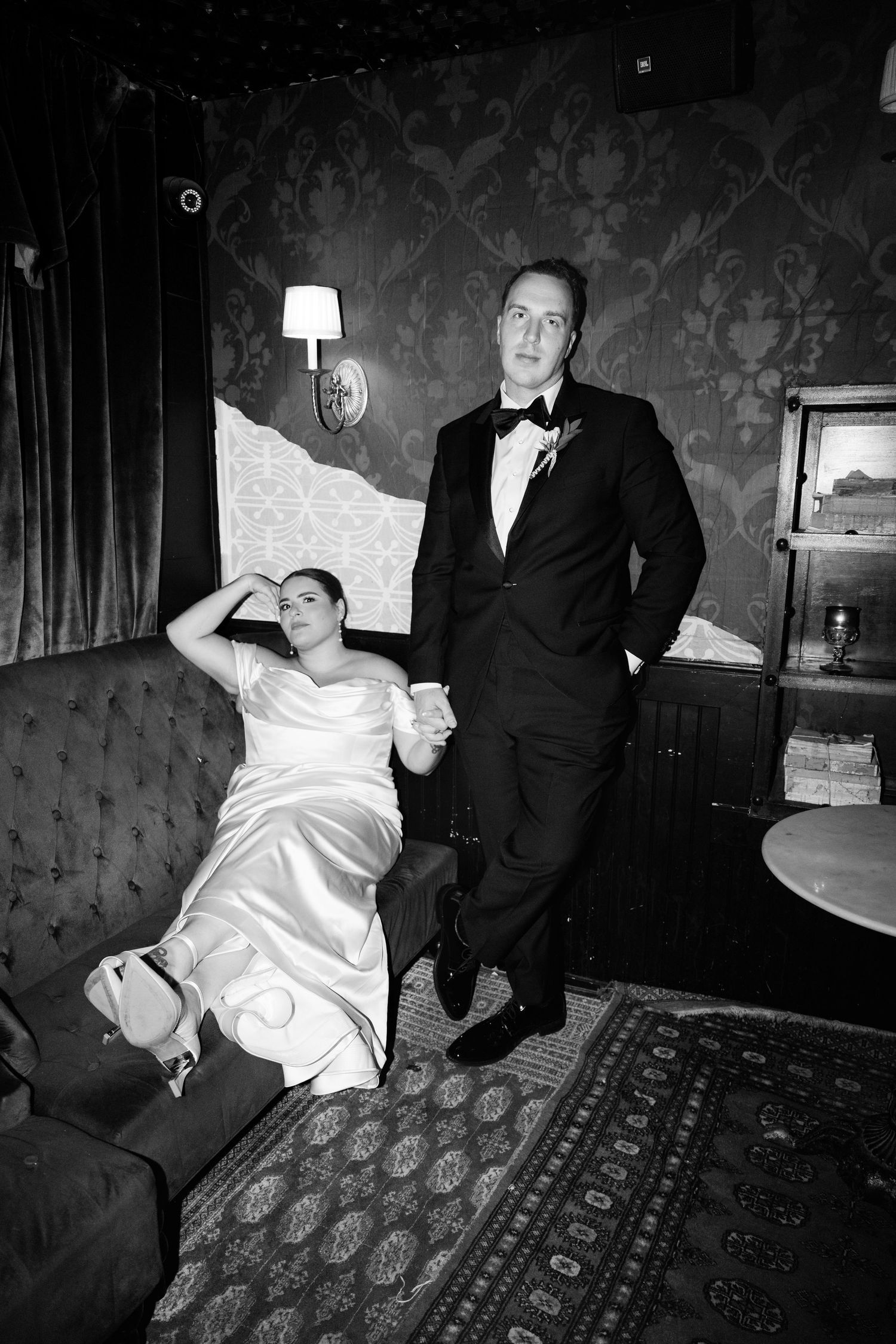 Black and white photo of bride reclining on sofa while groom stands holding her hand.