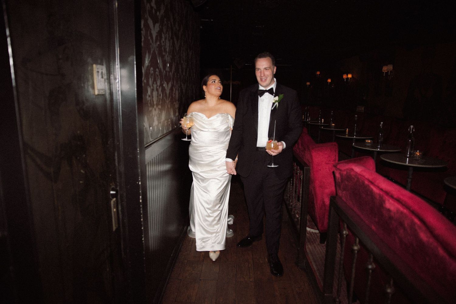 Bride and groom walk together holding champagne glasses in dimly lit elegant venue corridor.