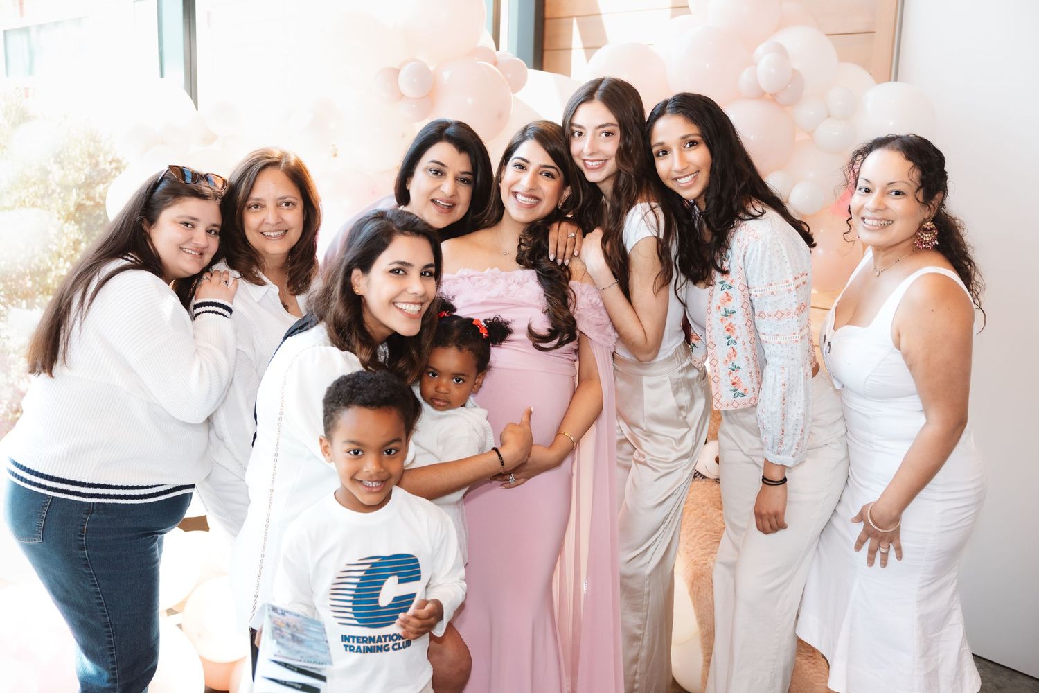 Group of women and children pose with pregnant woman in pink dress at baby shower.