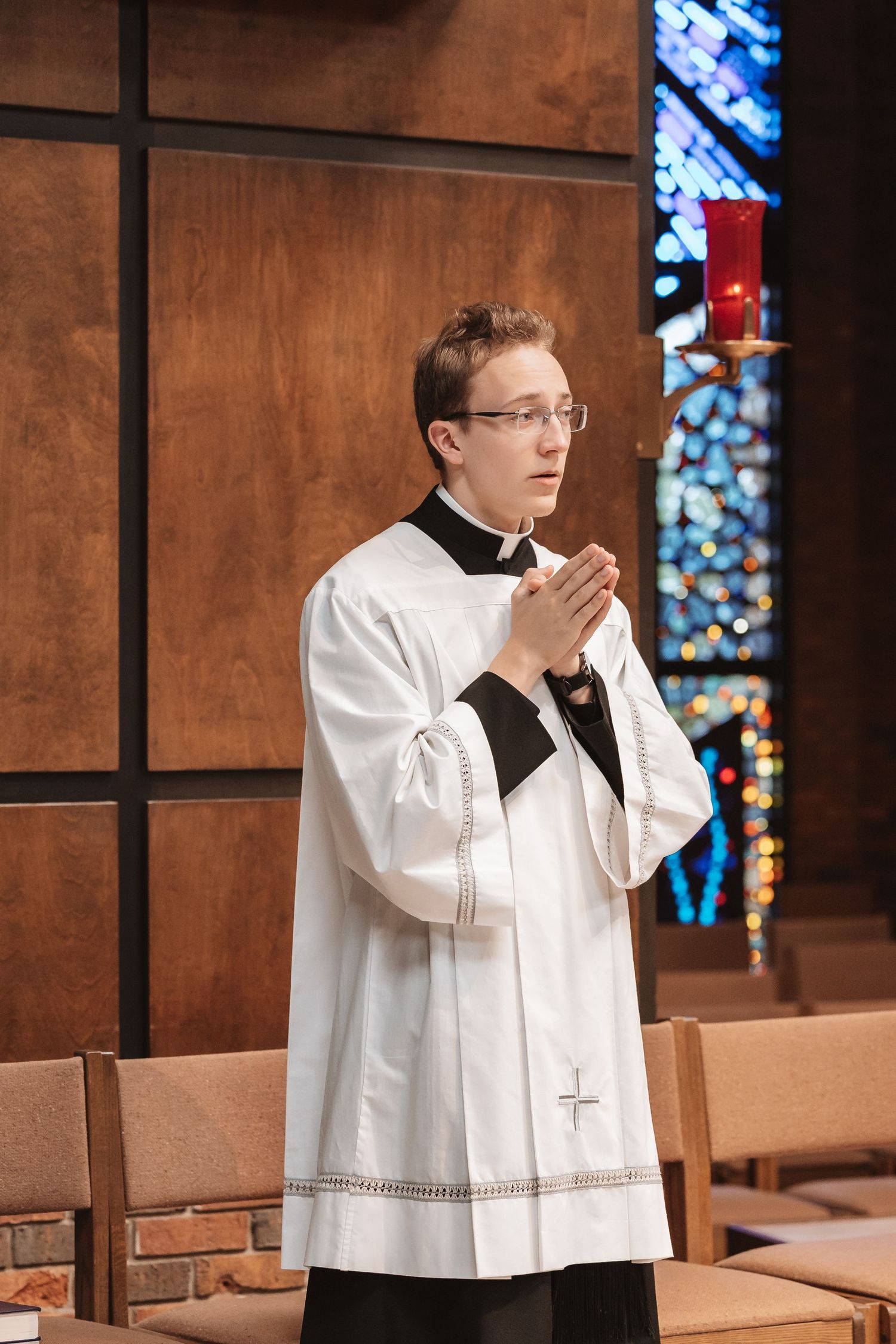 Young altar server in white vestments prays with hands clasped in church with stained glass window.
