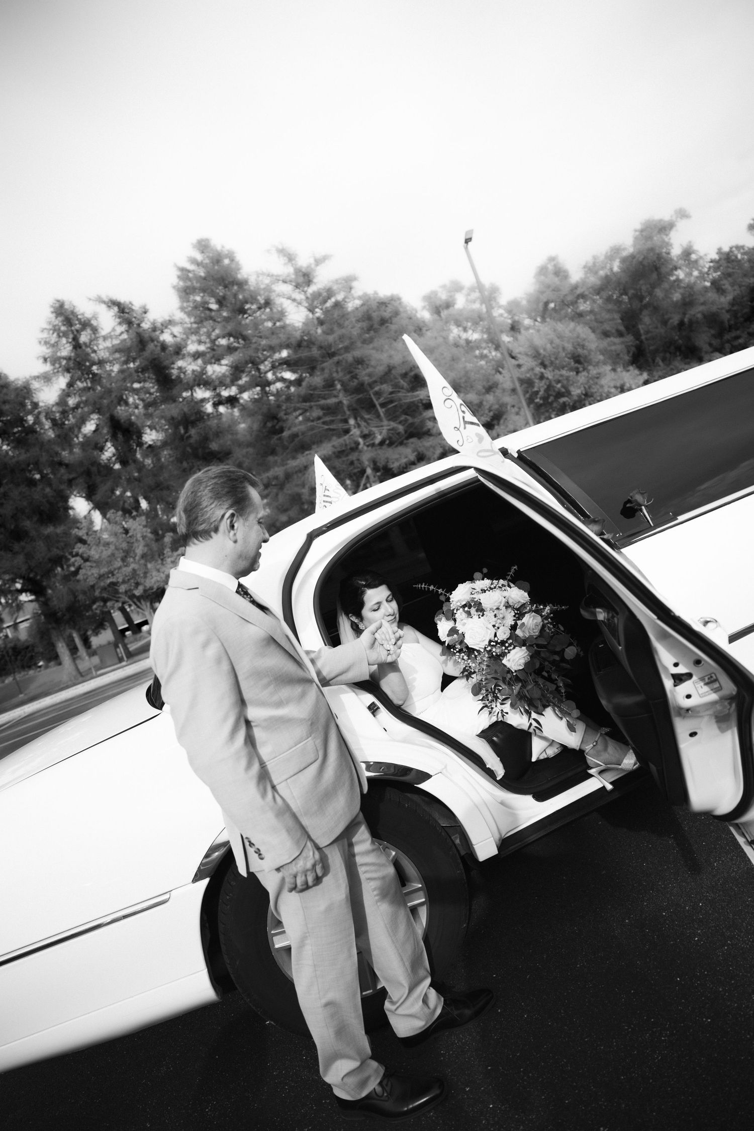 Groom helps bride exit white wedding limousine while she holds a floral bouquet, black and white photo.