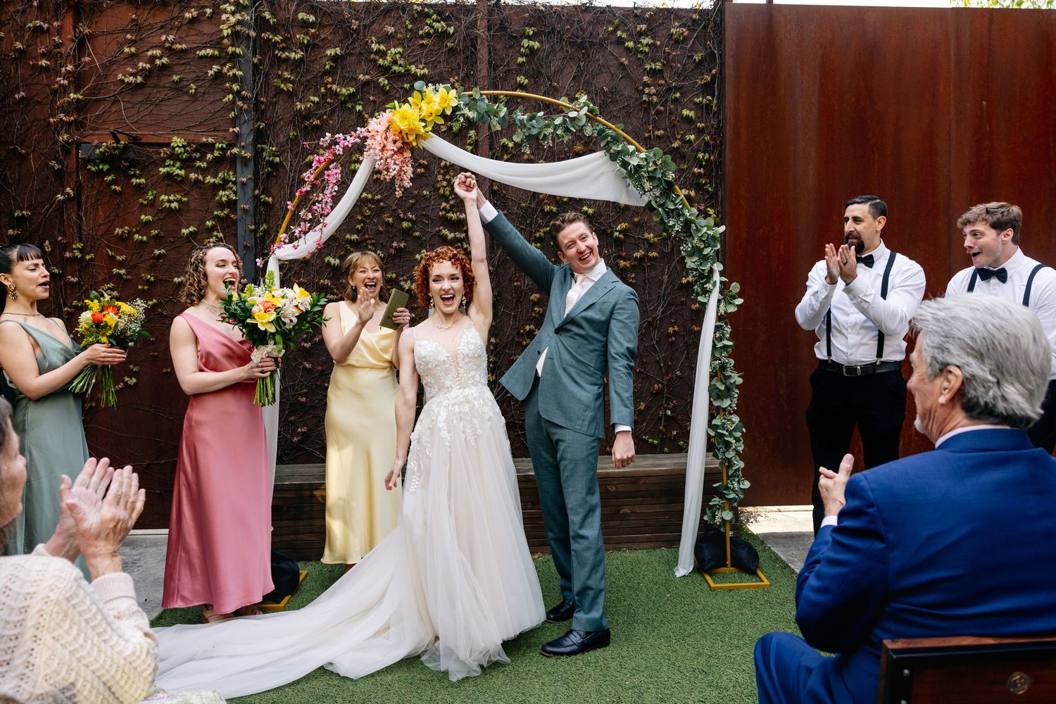 Bride and groom celebrate joyfully under a floral arch surrounded by colorful bridesmaids and groomsmen at an outdoor wedding.