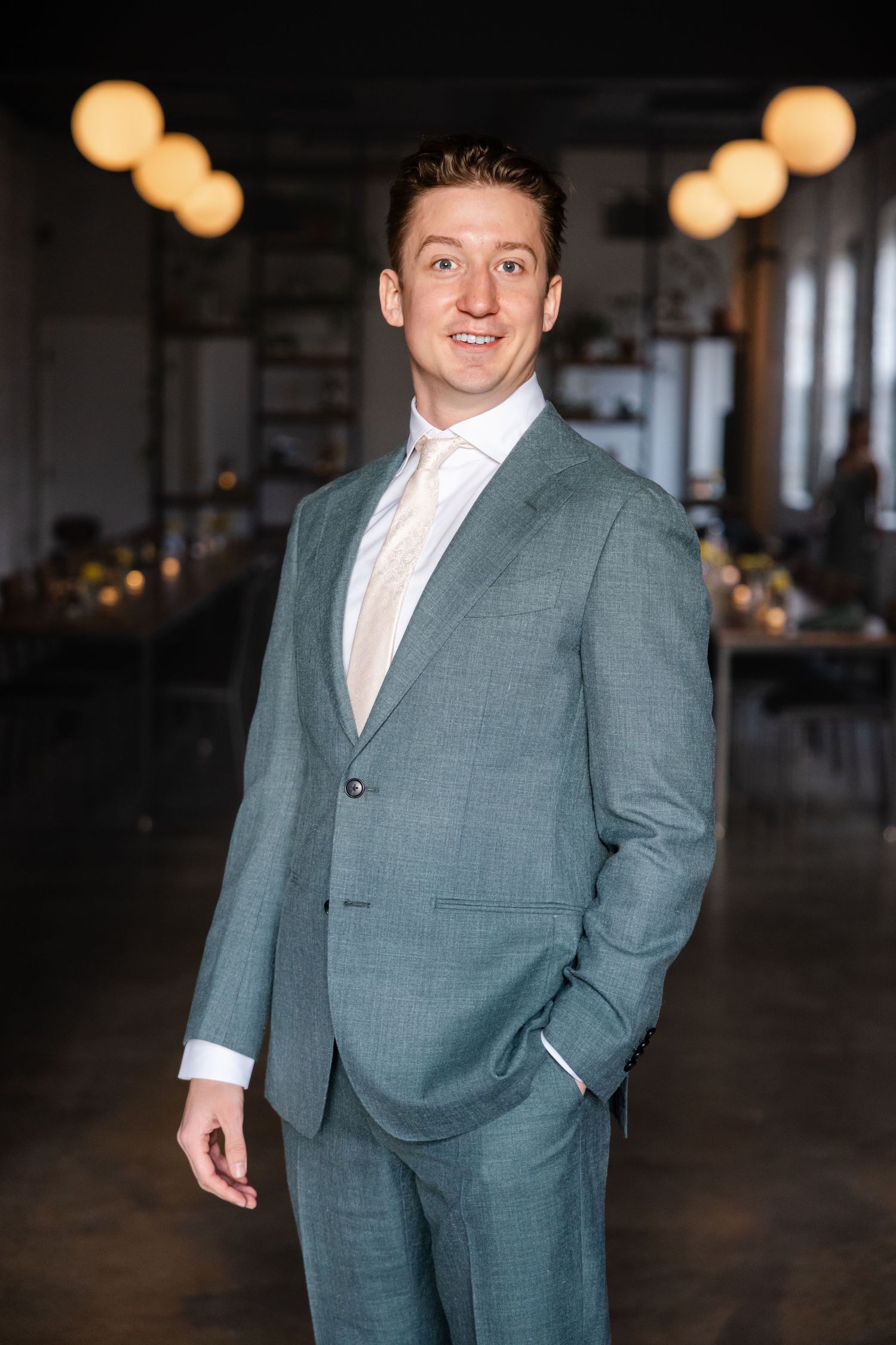 Handsome groom in teal suit smiles confidently in elegantly decorated wedding reception venue.