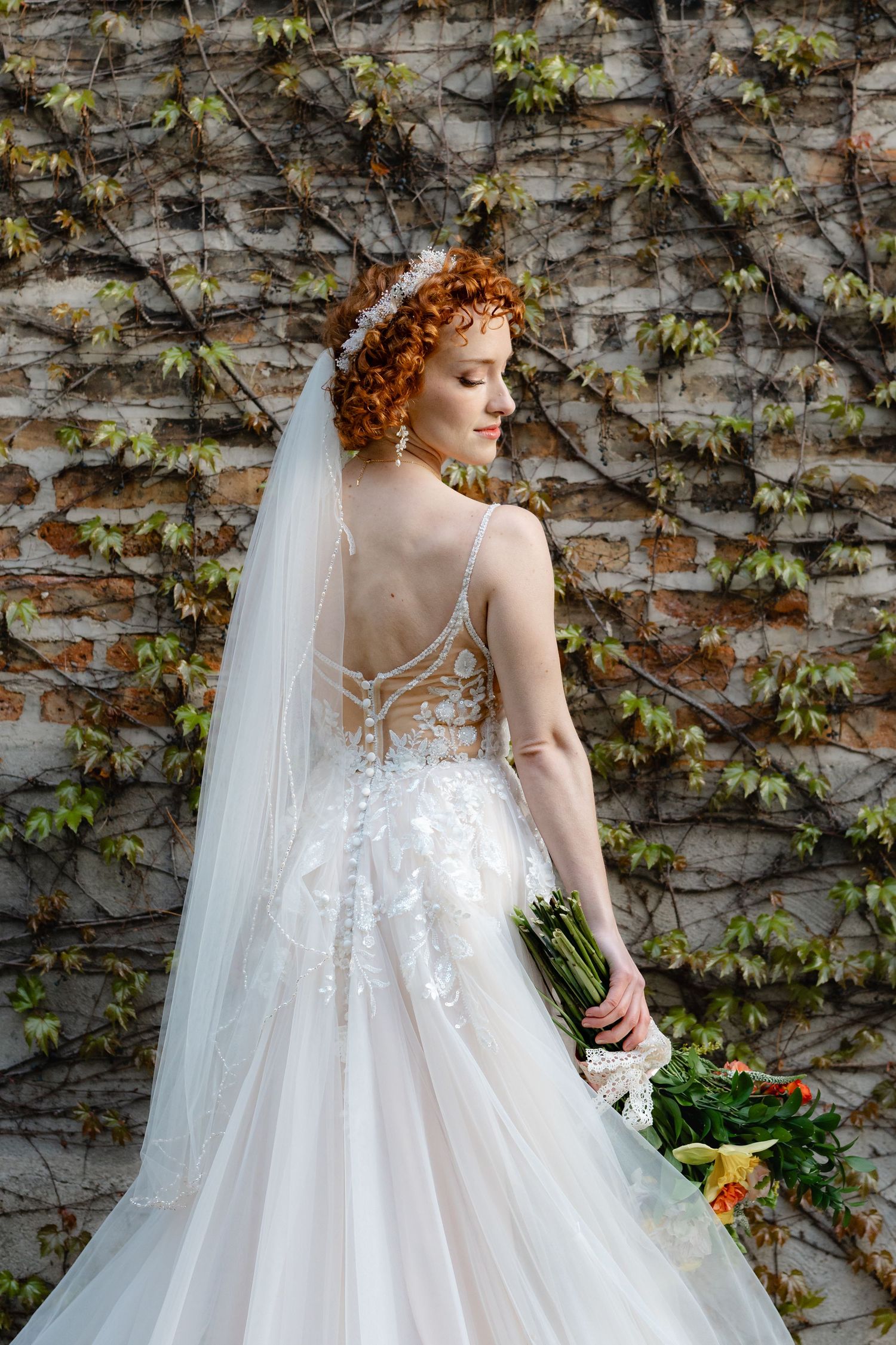 Redhead bride in backless lace wedding gown with veil holds bouquet before ivy-covered brick wall.