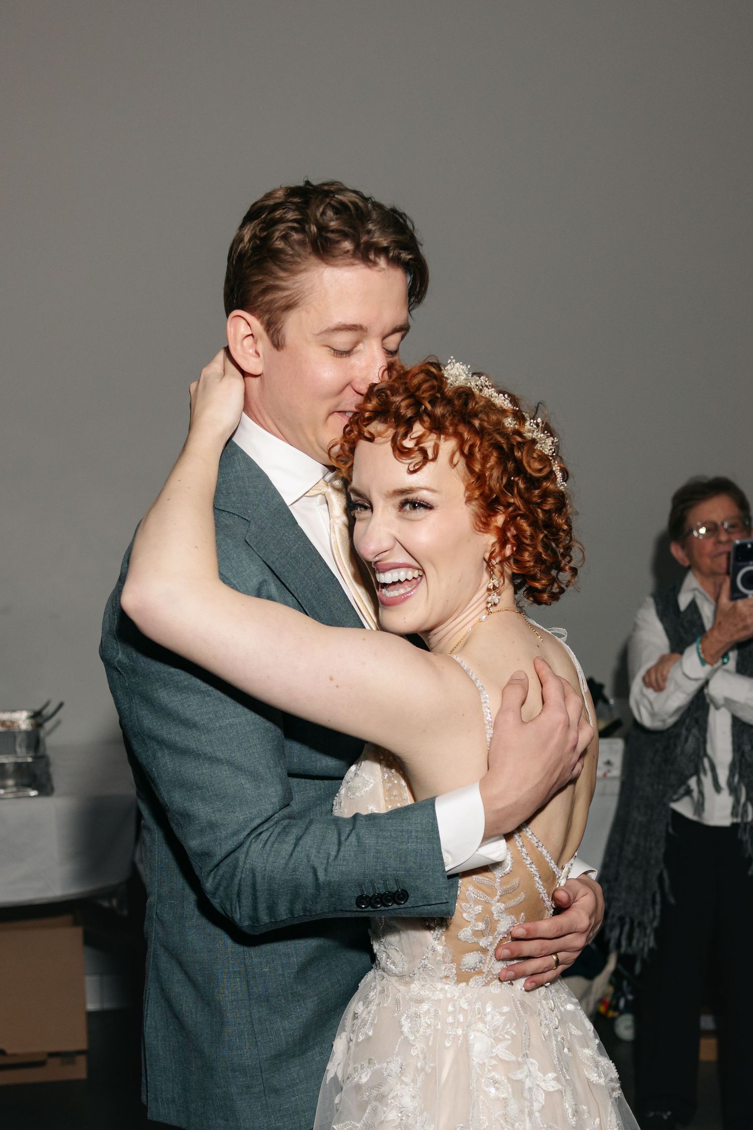 Joyful redhead bride in lace gown laughs during first dance with groom in teal suit at wedding reception.
