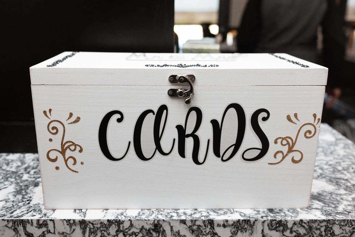 White wedding card box with black cursive lettering and gold floral accents on a decorated table.