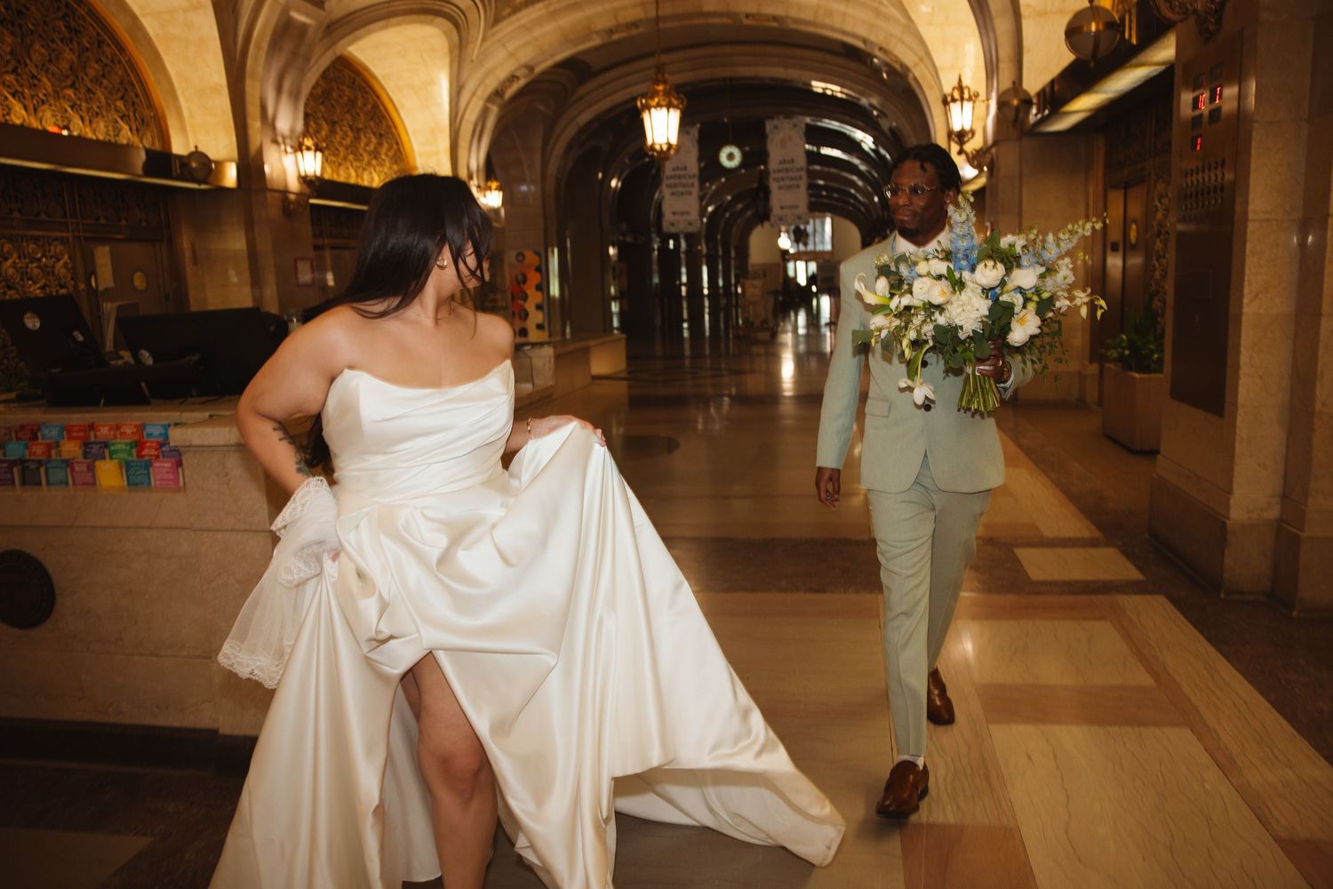 Bride and groom walking through elegant arched hallway, she holds dress while he carries white bouquet.