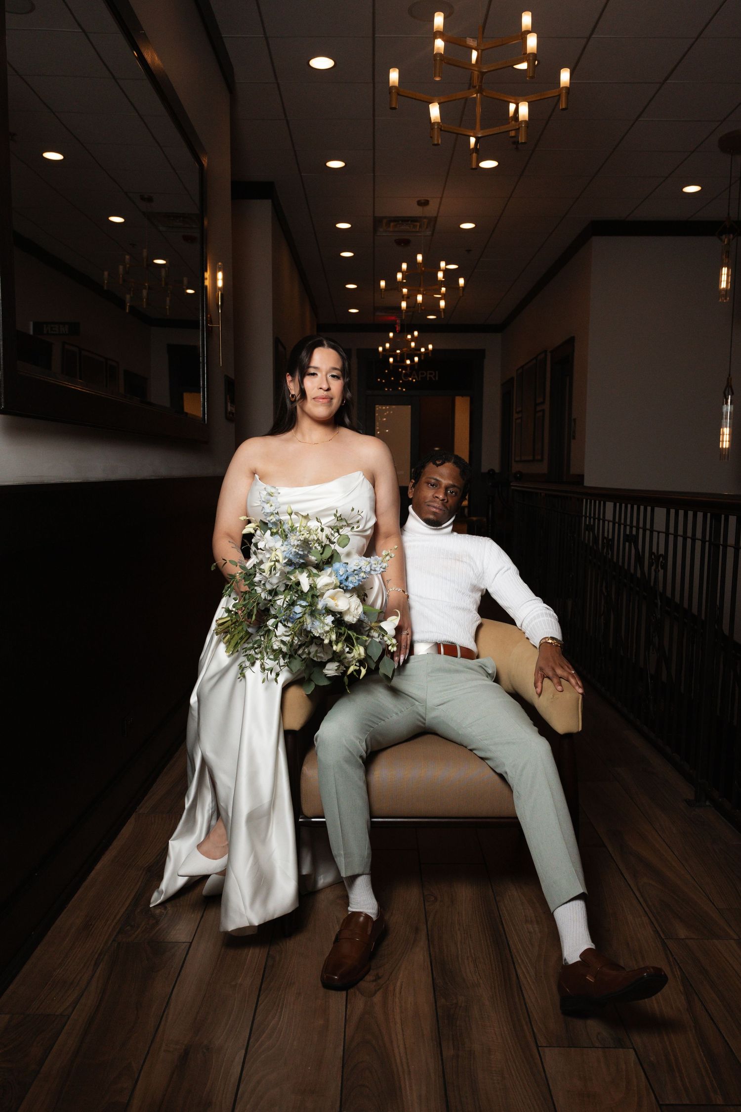 Bride holding blue floral bouquet stands beside seated groom in mint suit in dark elegant venue.