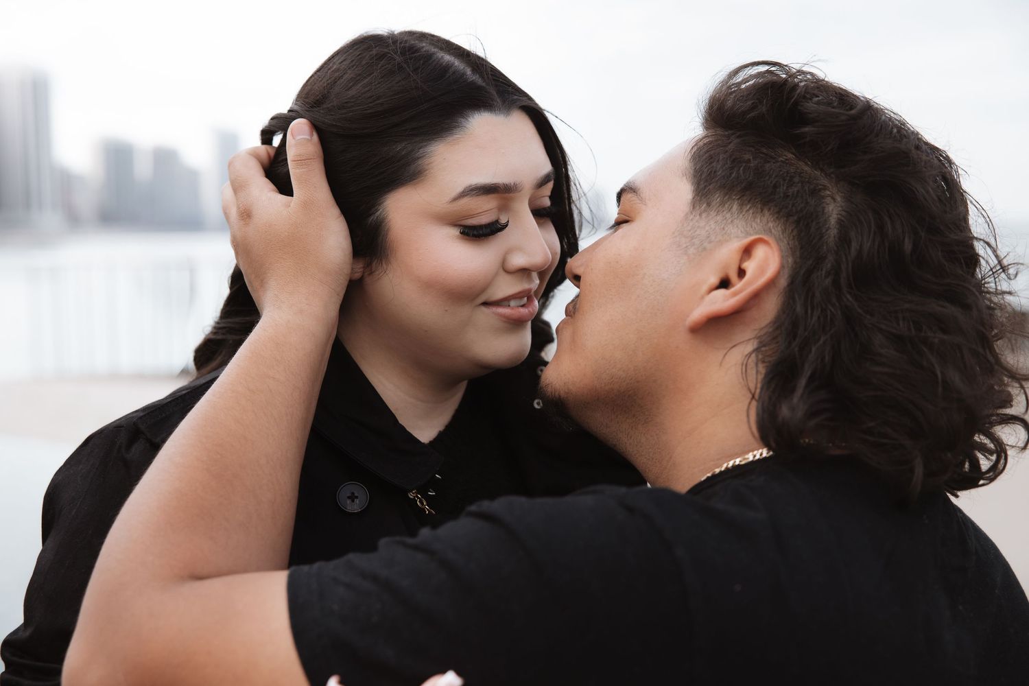 Romantic couple gazing at each other closely with a blurred waterfront city skyline in the background.