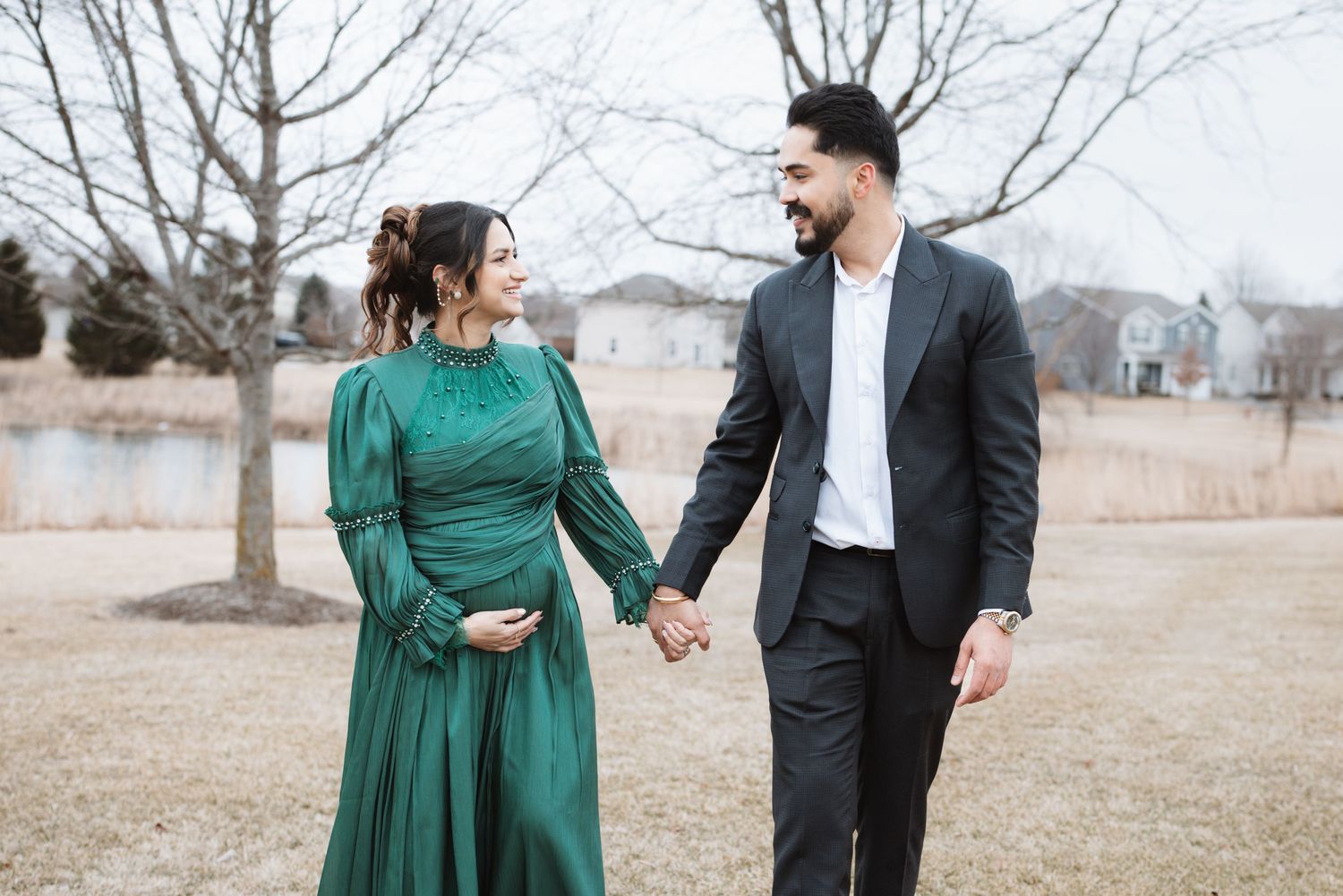 Expecting couple holding hands in green dress and black suit walking outdoors near lake.