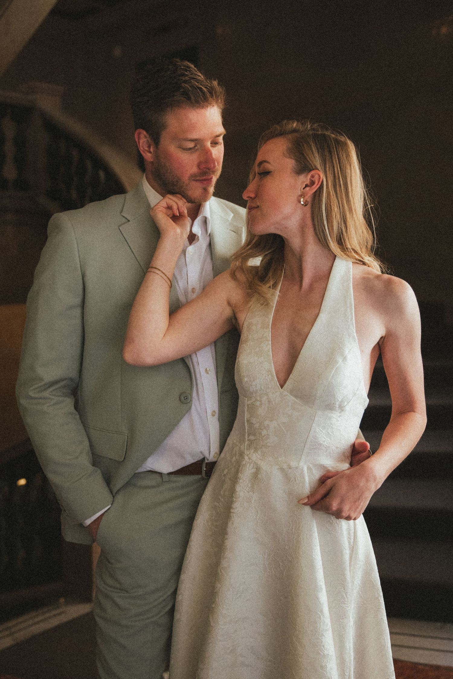Bride in white halter gown and groom in sage suit share an intimate moment indoors.