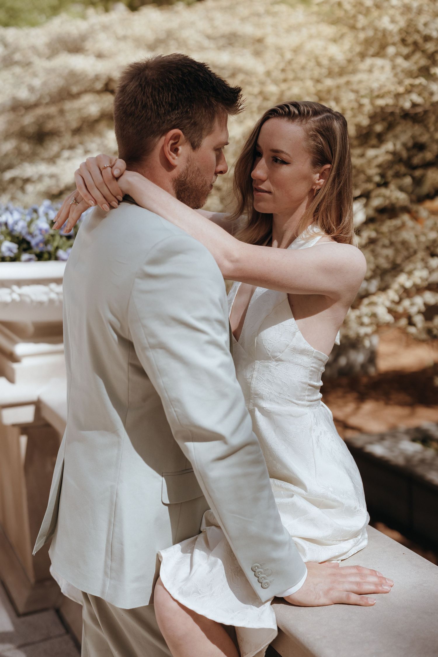 Bride in white halter gown embraces groom in beige suit on an outdoor terrace surrounded by greenery.