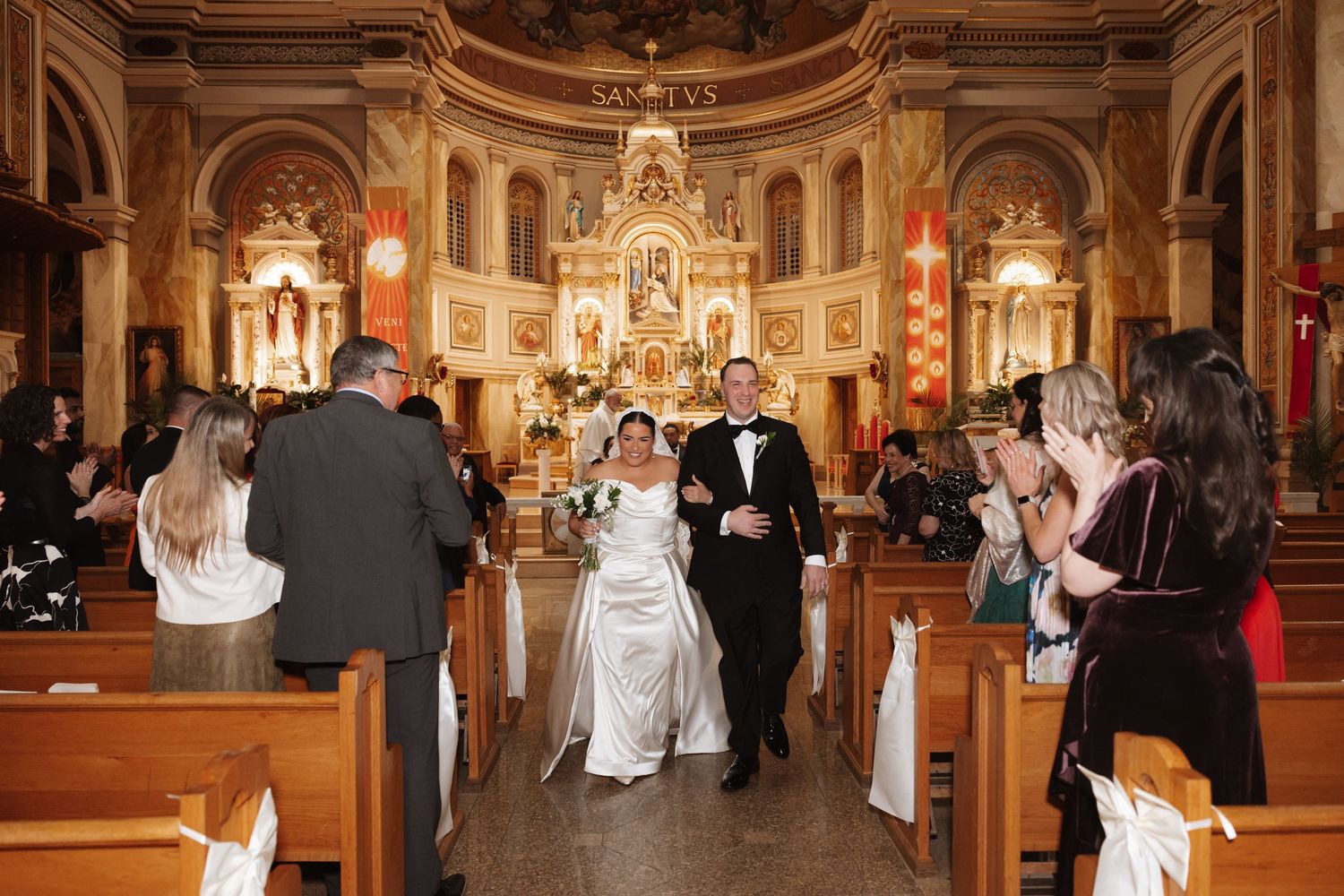 A bride and groom walk down the aisle of an ornate Catholic church surrounded by applauding wedding guests.