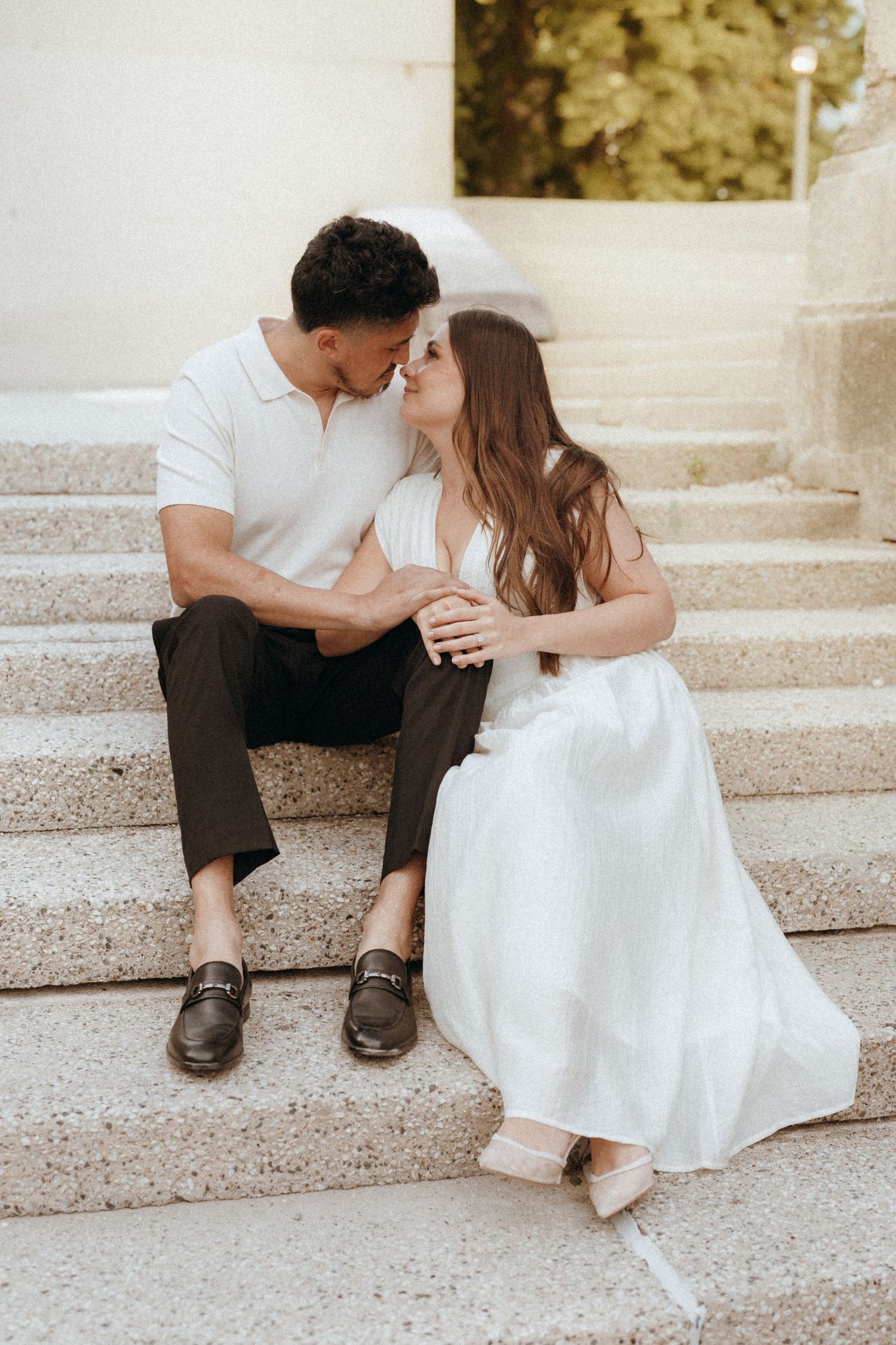 Engaged couple sits closely on stone steps, sharing an intimate moment in a white dress and polo shirt.