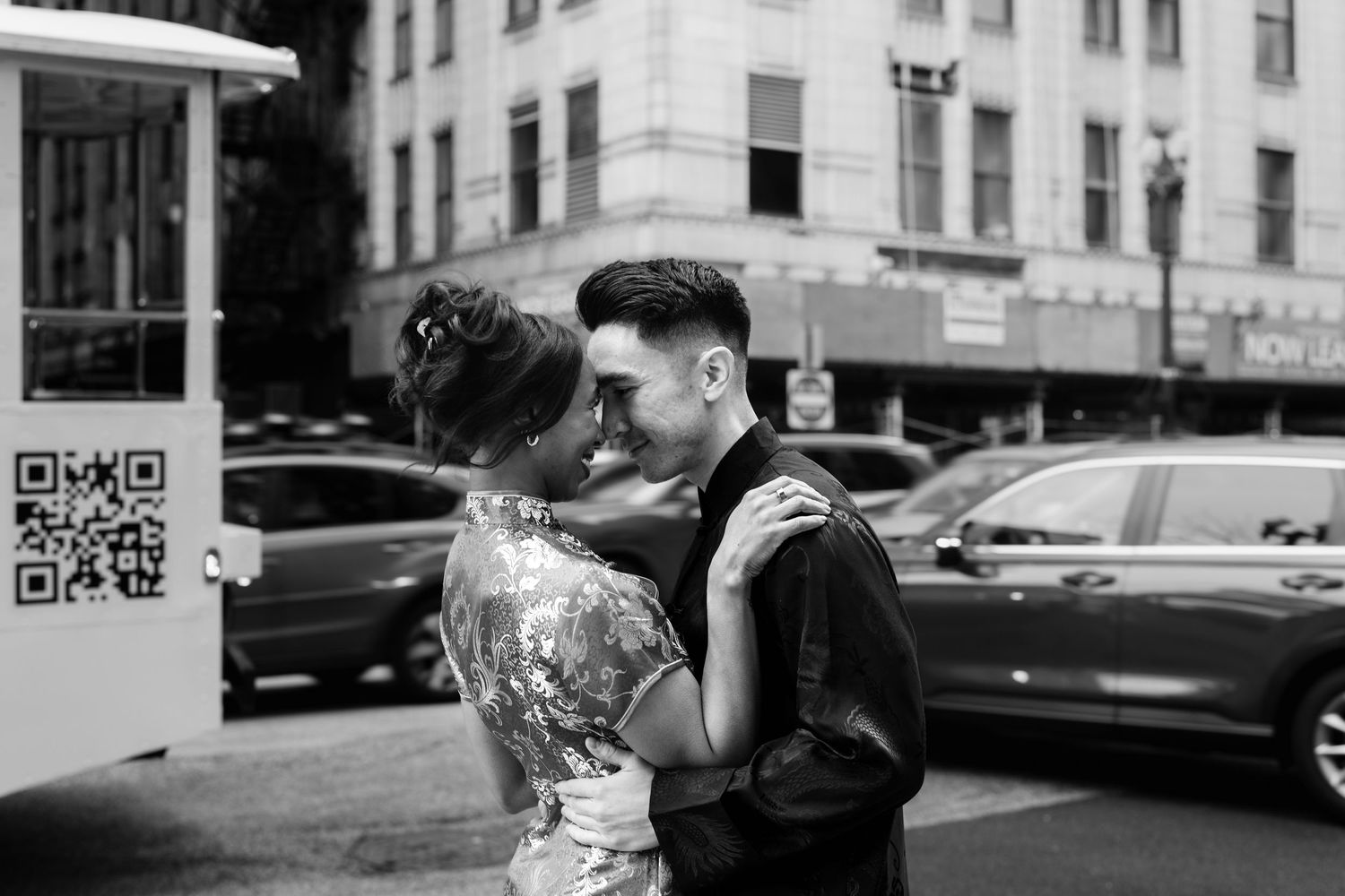 Black and white photo of couple in Chinese traditional attire sharing a kiss on a bustling city street.