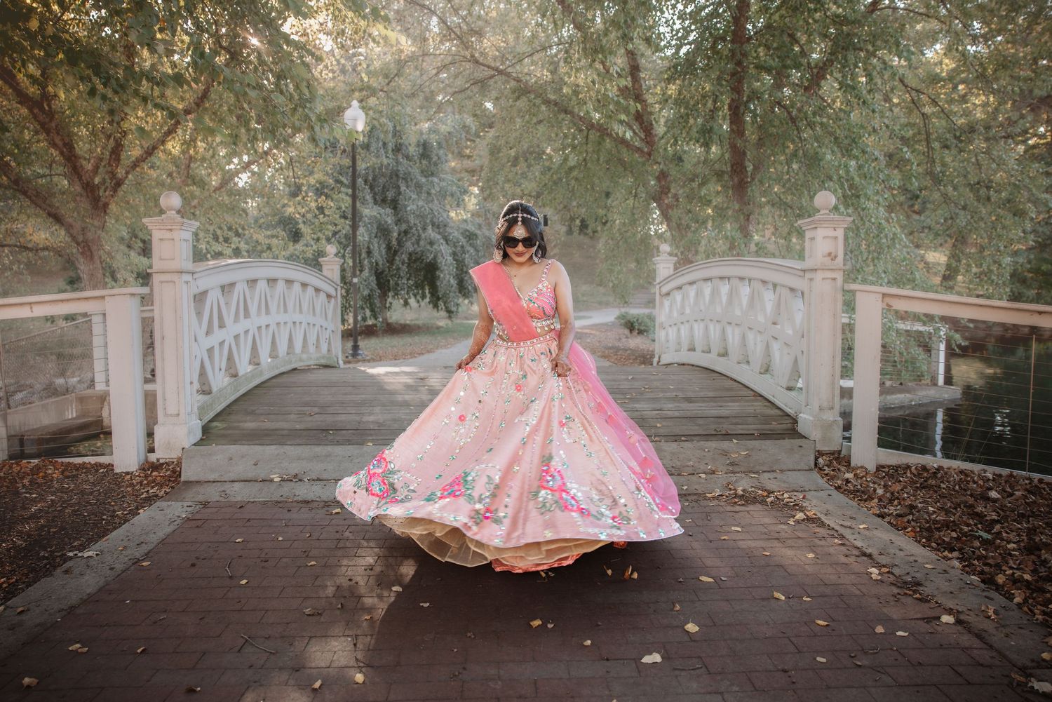 Bride twirling in pink floral lehenga on wooden bridge in lush garden setting.