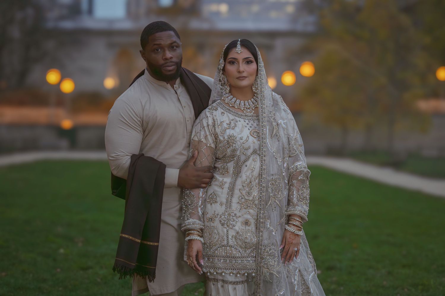 Groom in cream kurta and bride in ornate white lehenga stand together in a park at twilight.