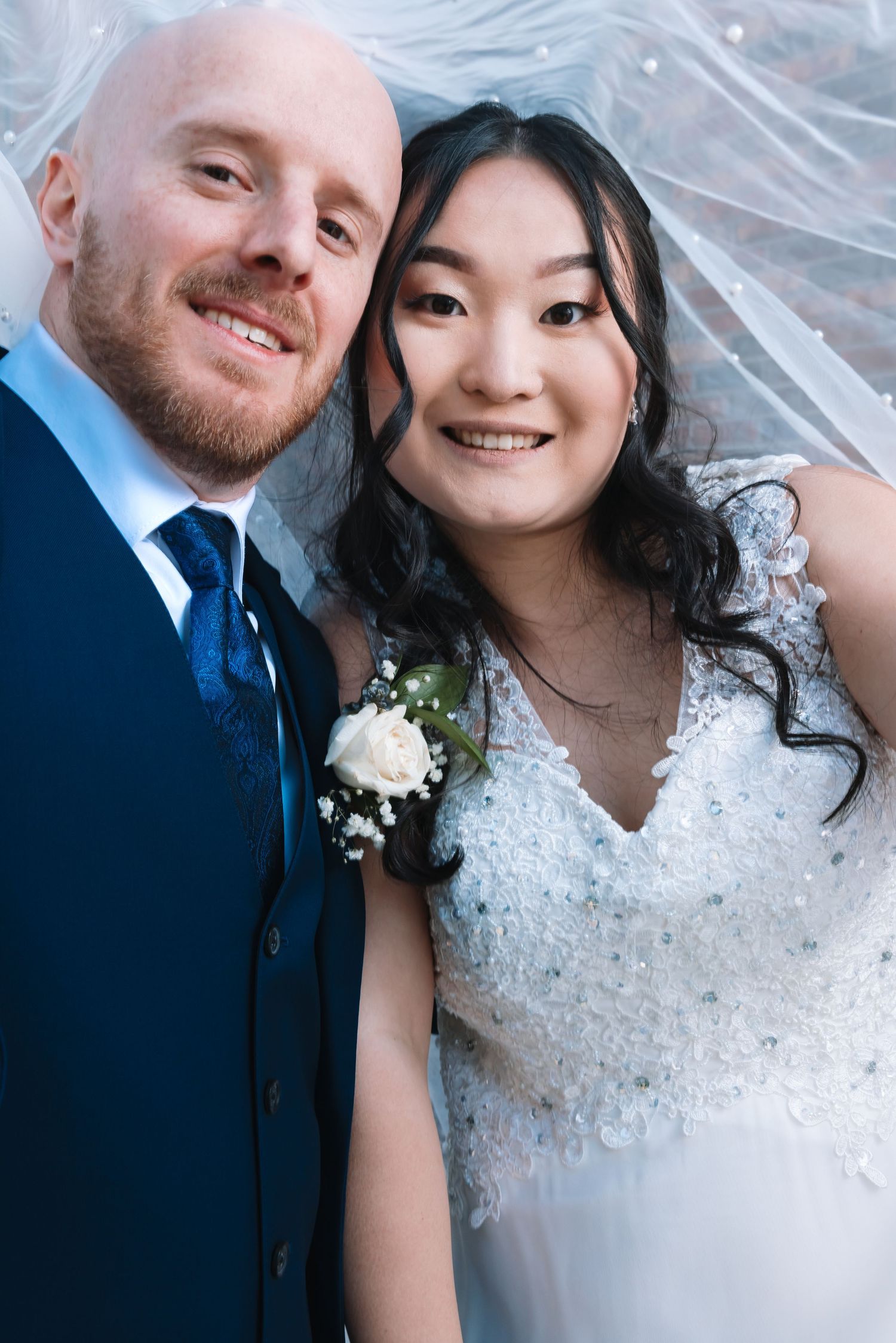 Smiling newlywed couple poses together under a pearl-adorned veil, groom wearing a boutonniere on wedding day.