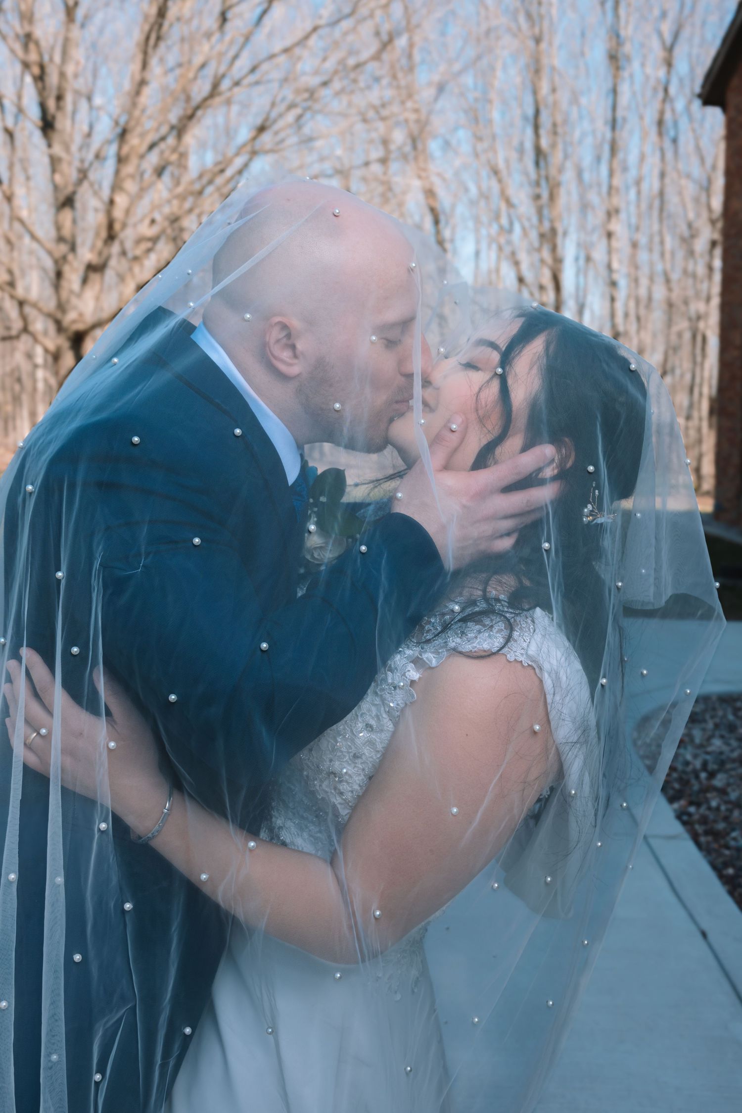 Wedding couple kissing passionately under a pearl-adorned veil with bare winter trees in the background.