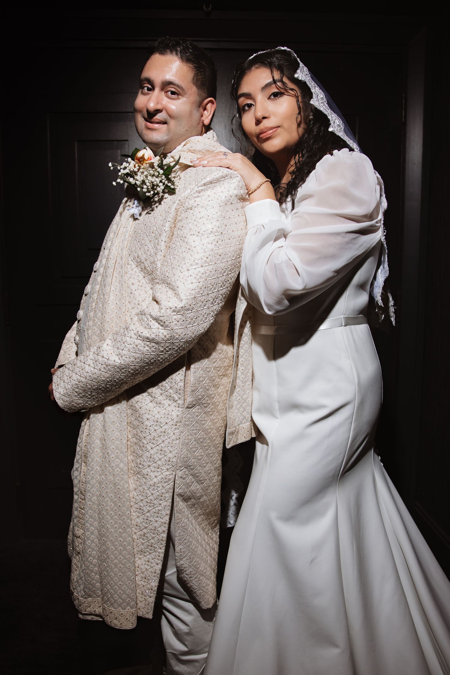 Bride in white dress leans on groom in cream sherwani, both smiling in a dark setting.