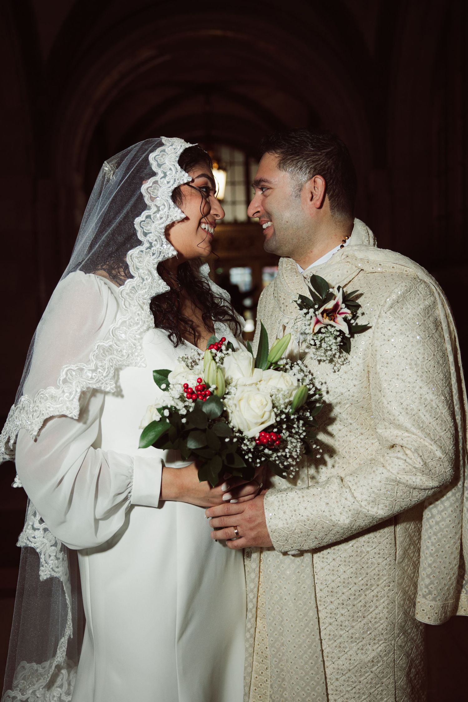 Smiling bride and groom holding a white floral bouquet inside a stunning arched corridor.