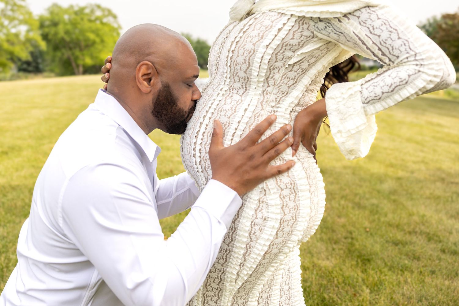 A man in white kisses a pregnant woman's belly outdoors.