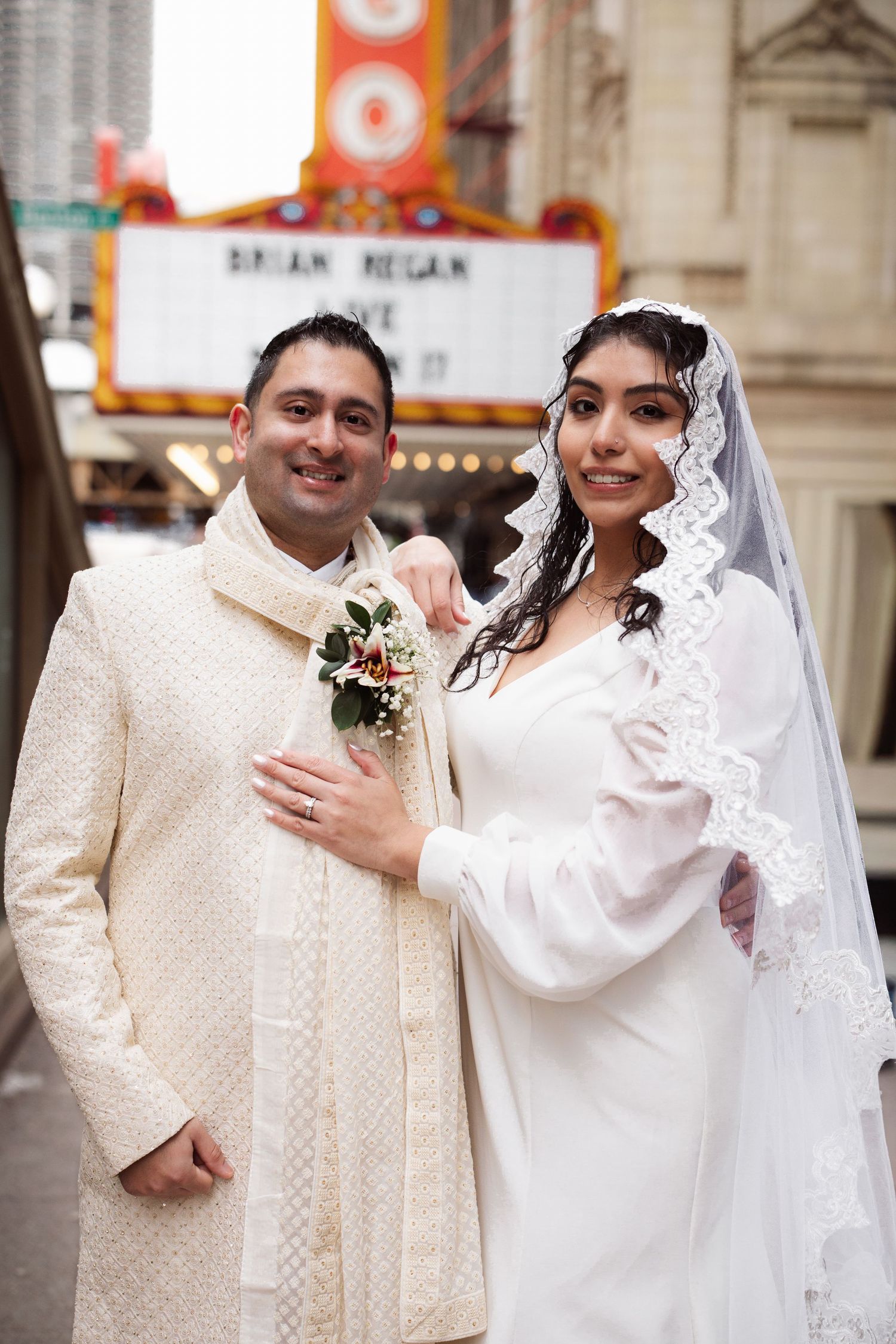 Happy bride and groom in white wedding attire pose outside Chicago Theatre marquee sign.