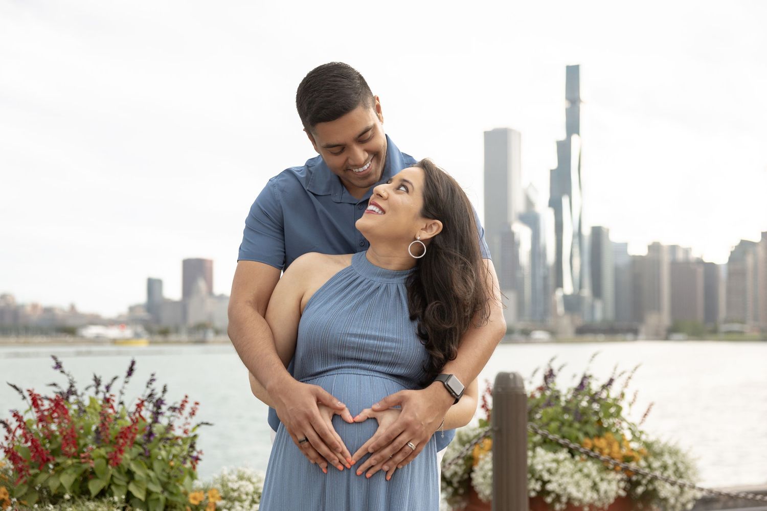 Happy expectant couple making heart shape over baby bump with Chicago skyline and waterfront behind them.
