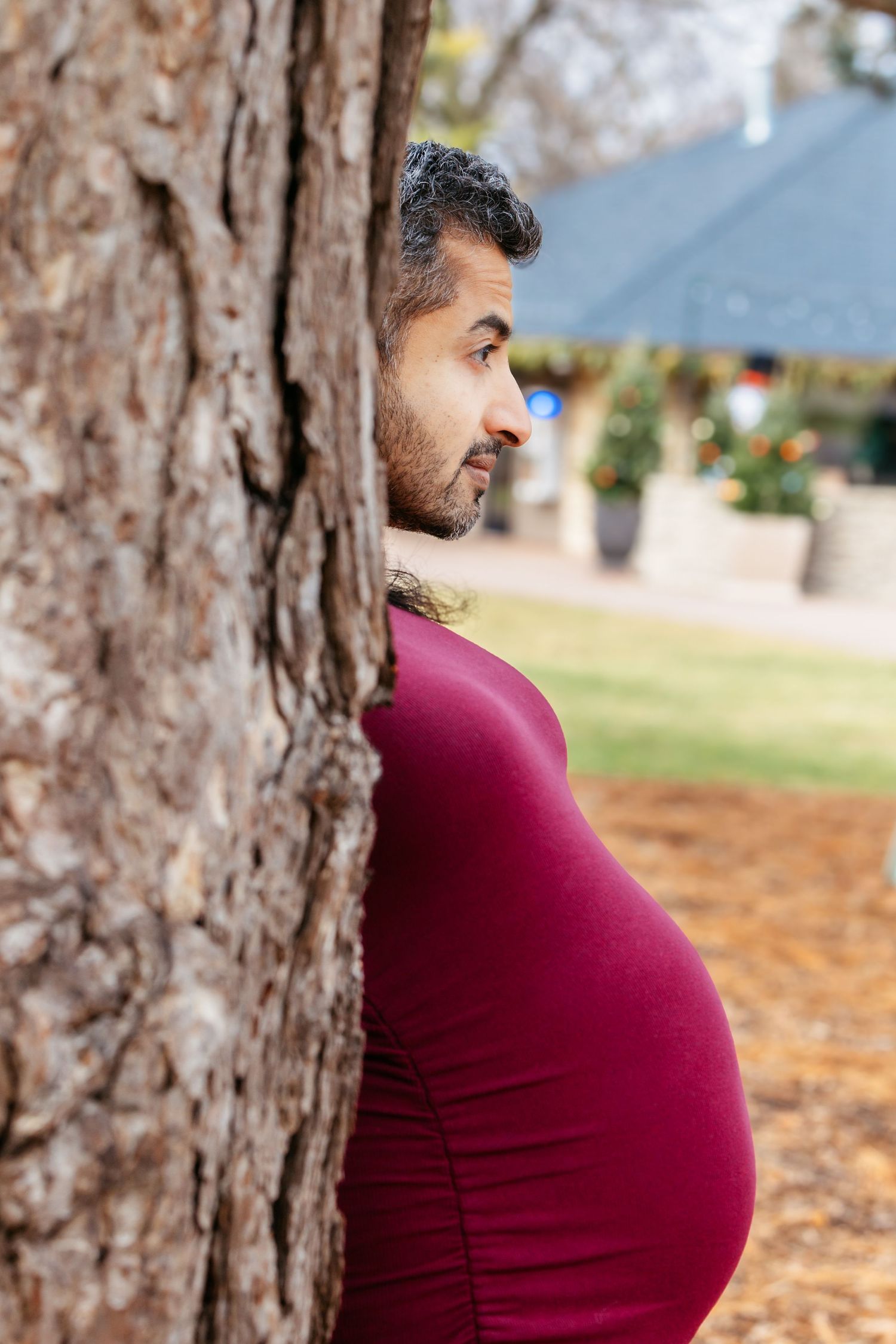 Pregnant woman in burgundy dress leaning against tree trunk outdoors.