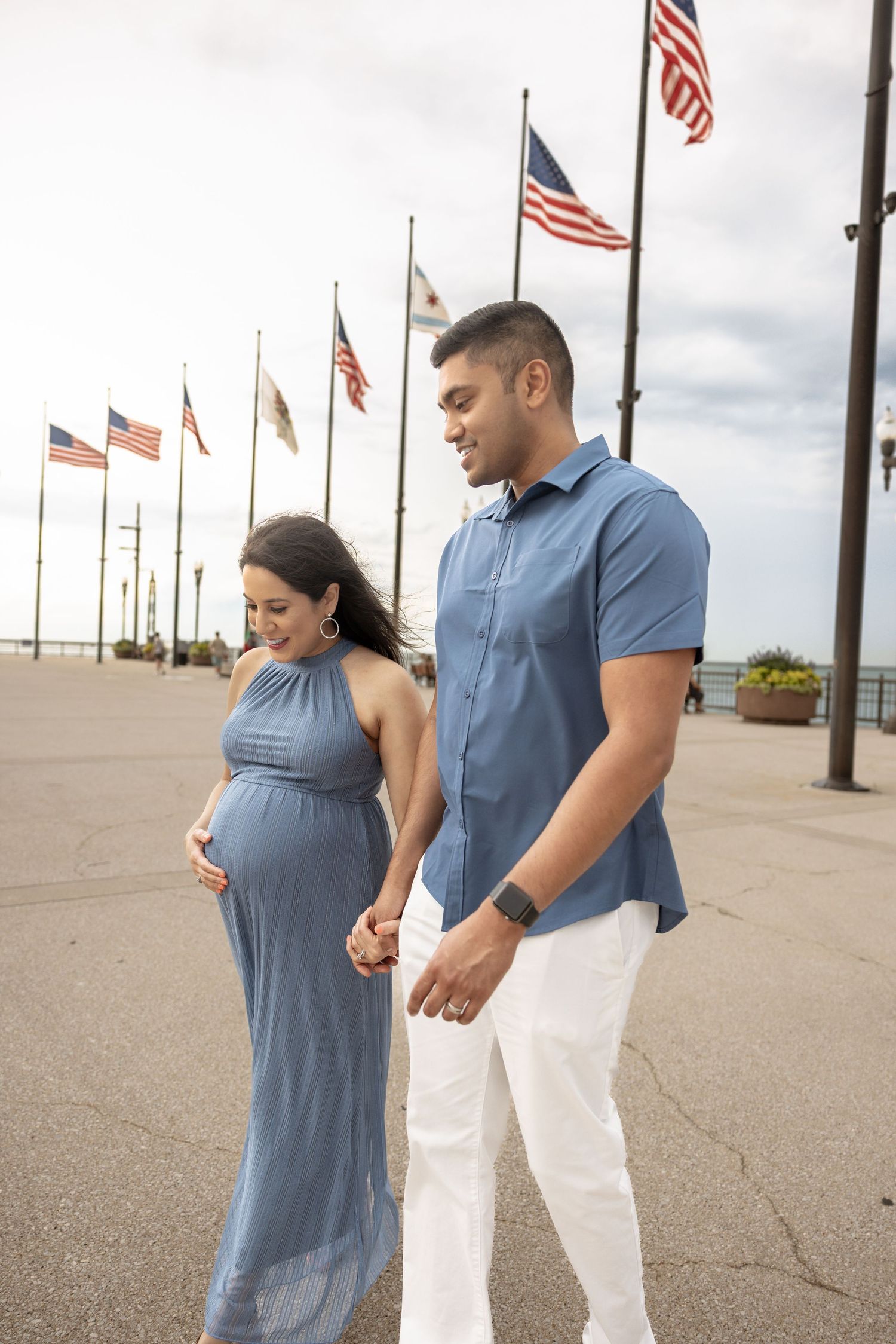 Pregnant couple holding hands walking near American flags on waterfront promenade.