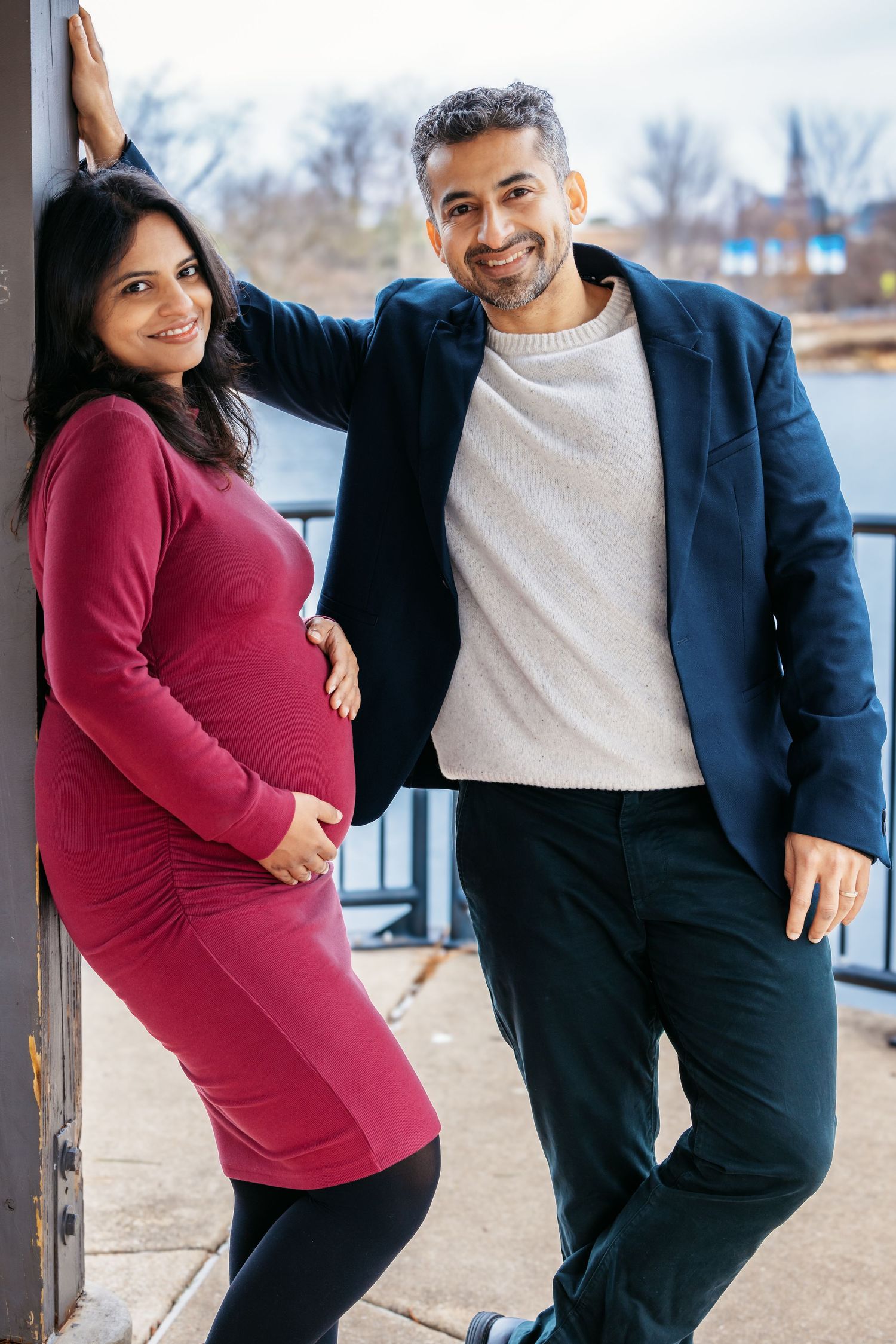 Joyful pregnant couple smiling together near a river waterfront railing.