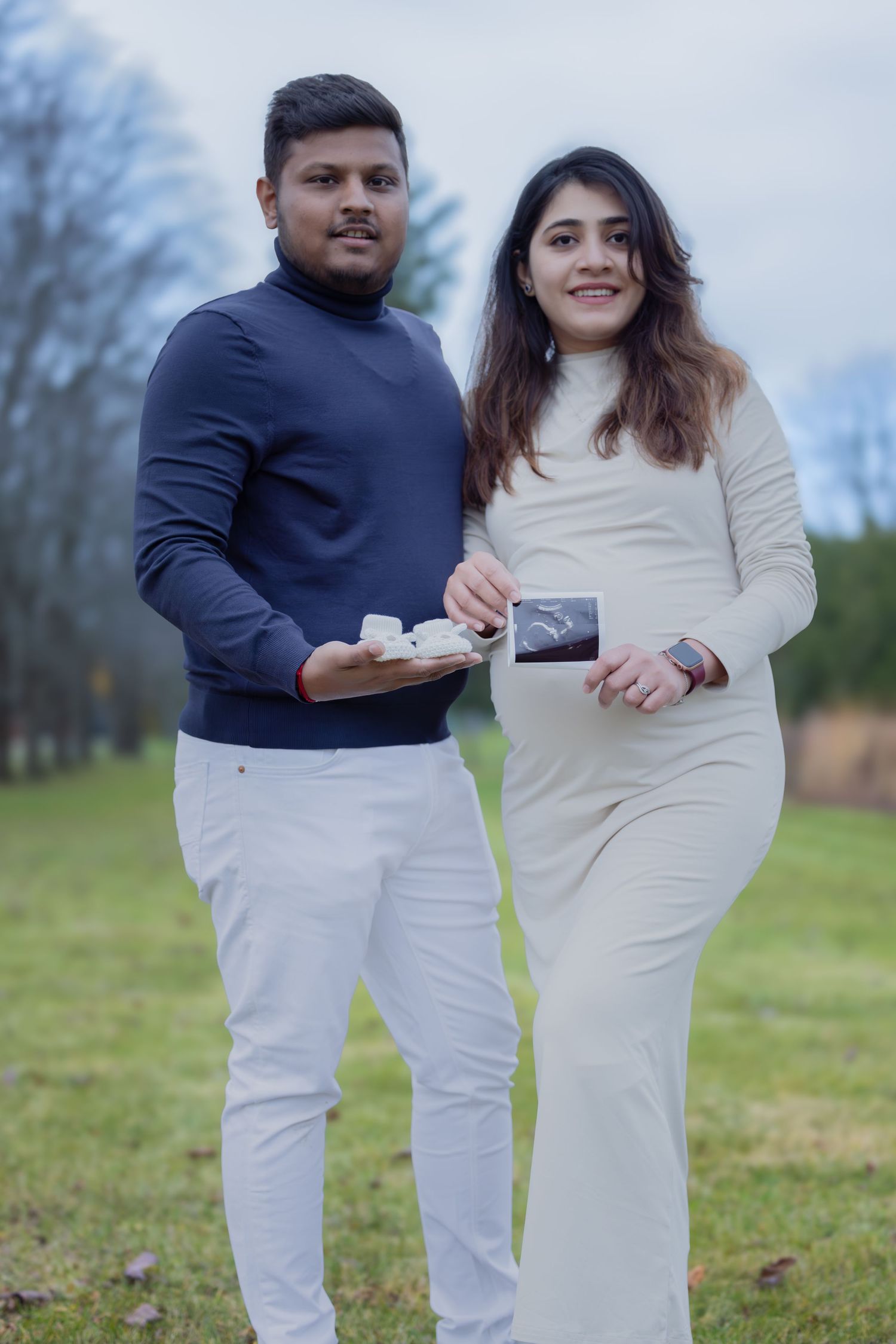 Smiling pregnant couple holding baby shoes and ultrasound photos outdoors.
