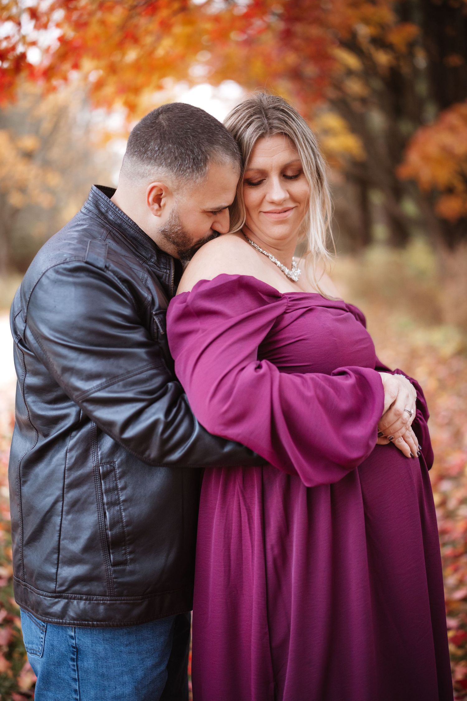 Man in leather jacket hugging pregnant woman in purple off-shoulder gown among fall foliage.