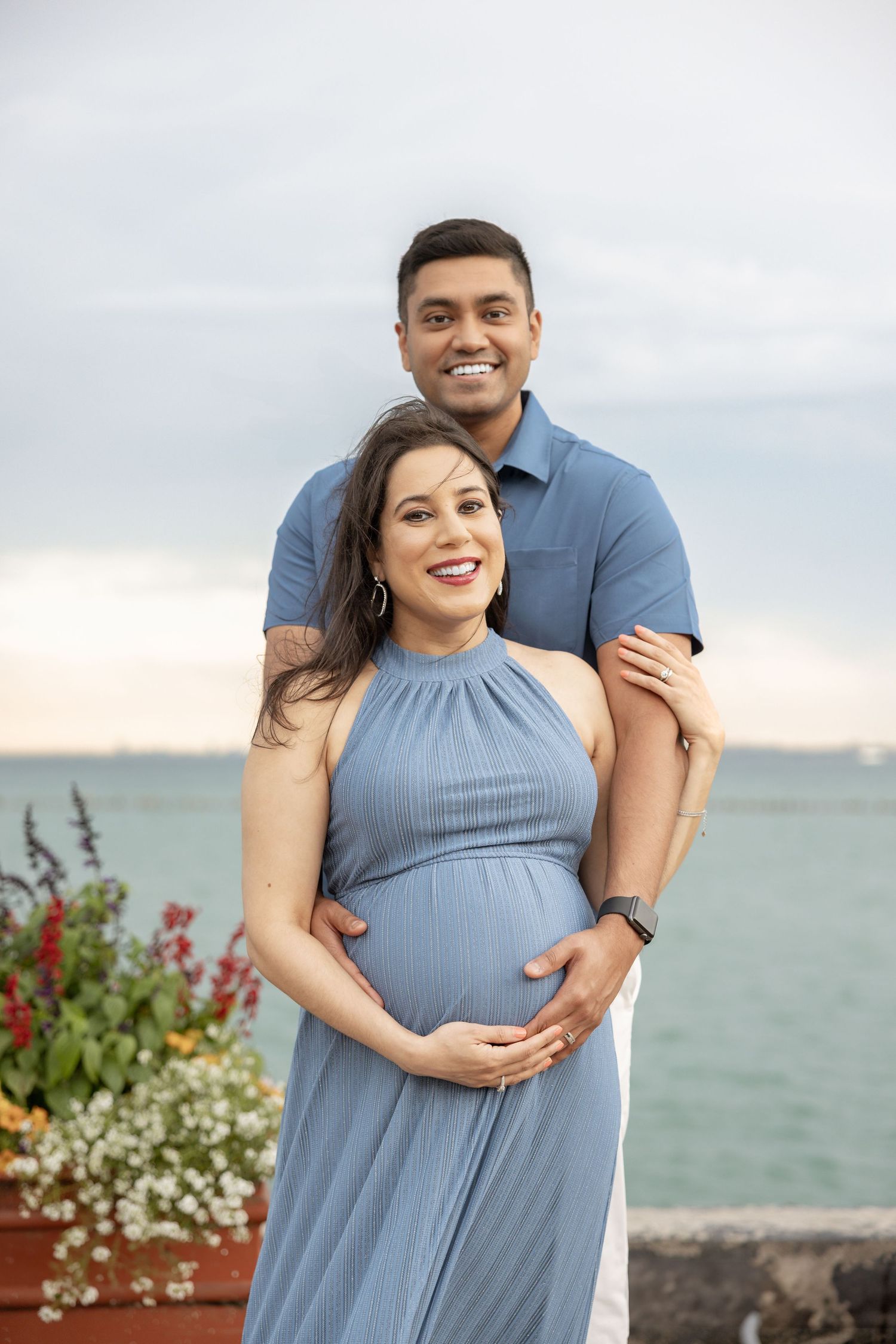 Joyful expecting couple posing outdoors by the water, woman in blue halter maternity dress cradling her baby bump.