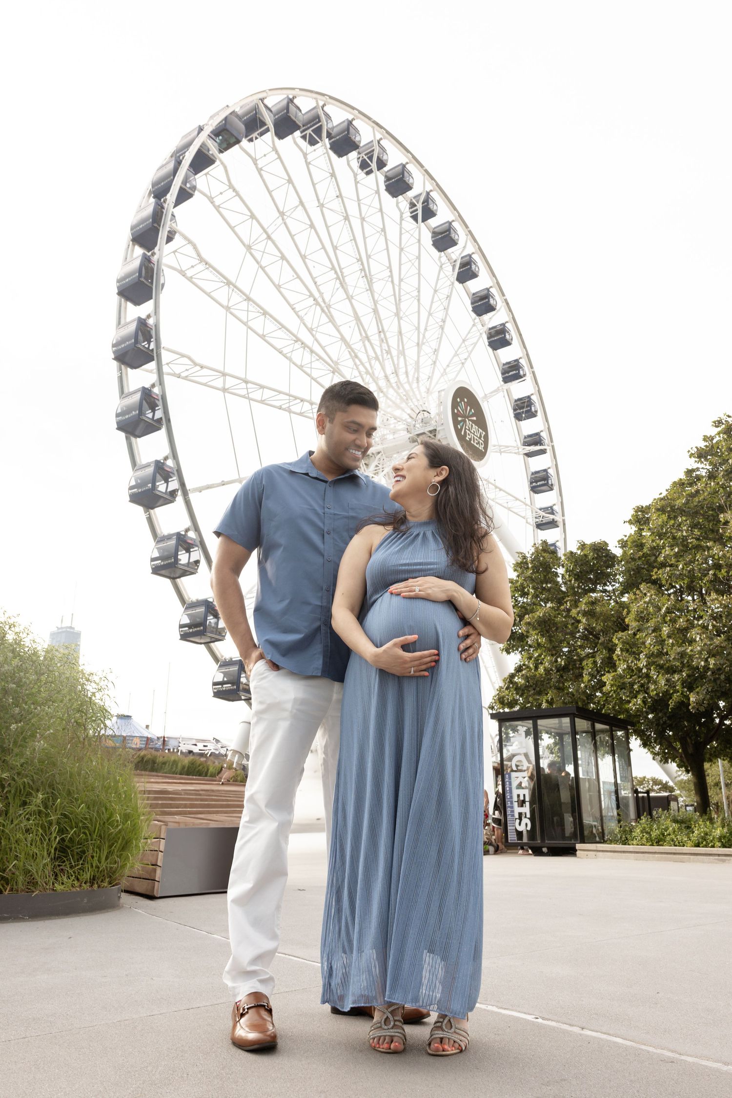 Pregnant couple standing before a large Ferris wheel, woman in blue maxi dress cradling her baby bump.